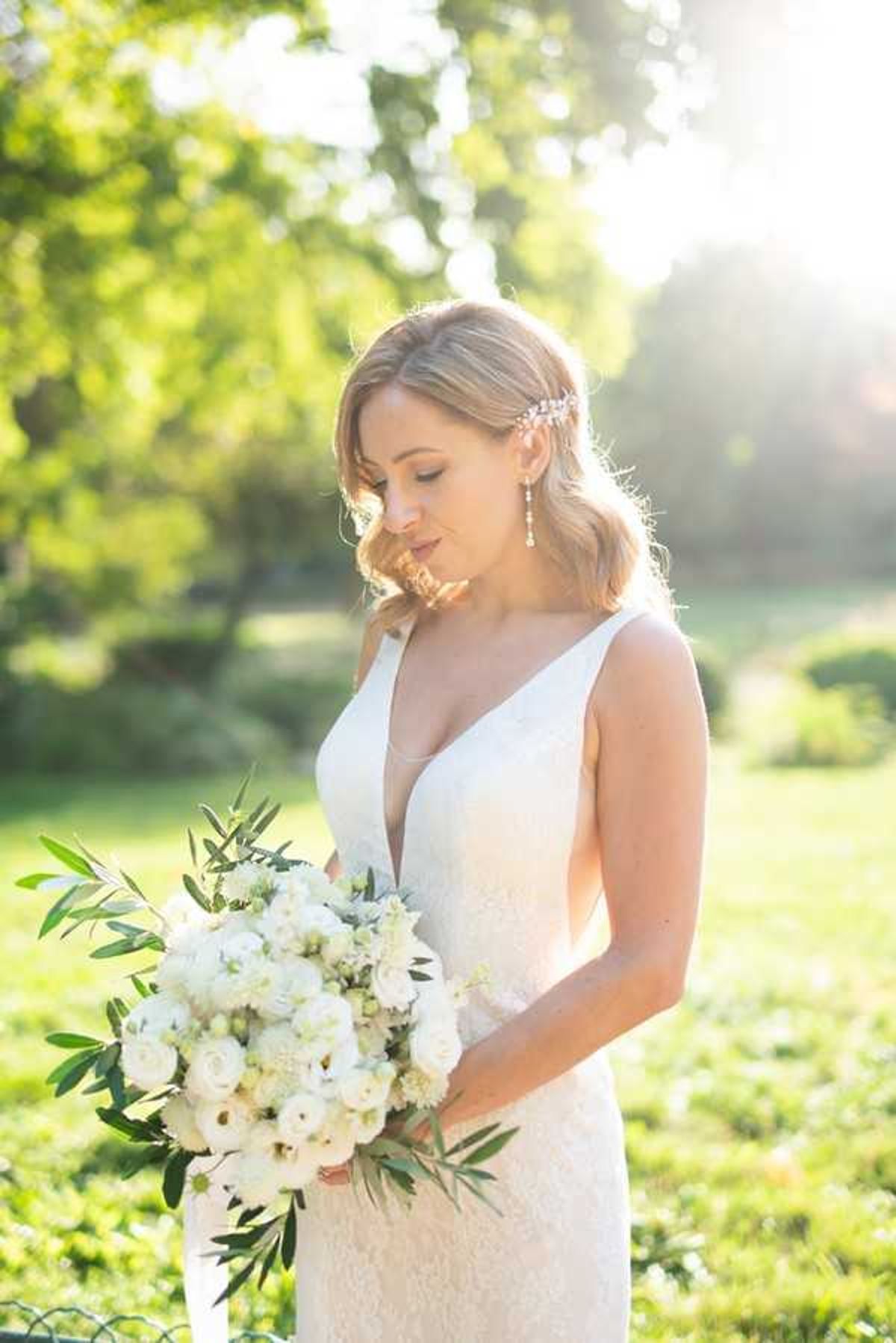 A bridal portrait taken outdoors in a garden setting, with warm golden backlight creating a sun-flare effect behind the subject. The bride wears a fitted ivory lace gown with a deep V-neckline and holds a rounded bouquet of white roses, ranunculus, and green olive branch foliage. Her blonde hair is worn in a loose half-up style with a delicate crystal or pearl hair accessory, complemented by drop earrings. She is looking downward with eyes closed in a posed, contemplative expression. The shot is a medium close-up portrait framing the bride from approximately the waist up.