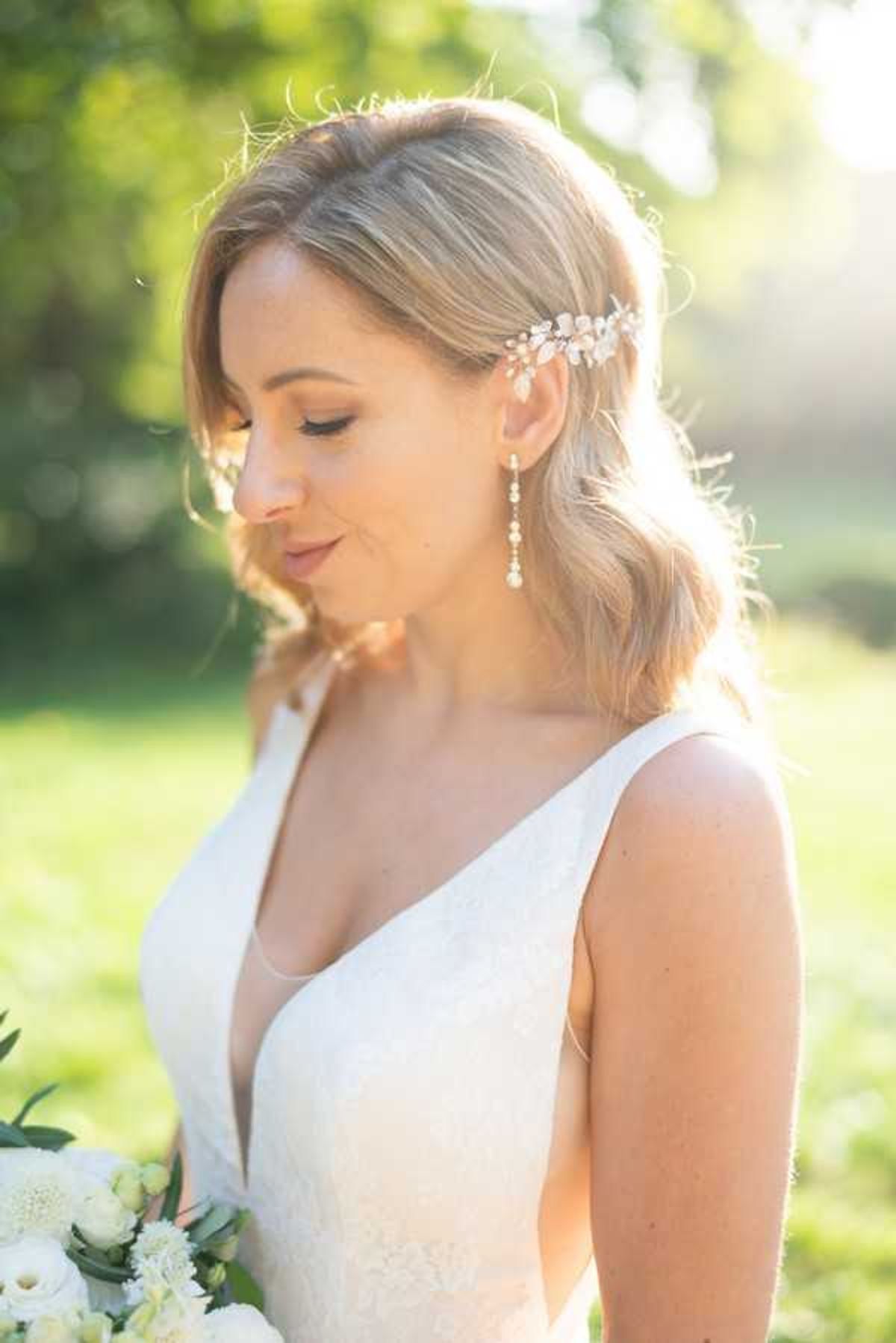 A close-up portrait of a bride shot outdoors in soft natural backlight. She wears a white lace gown with a deep V-neckline, long pearl and crystal drop earrings, and a delicate floral hair comb positioned at the side of her loosely waved blonde hair. She holds a bouquet of white ranunculus and ivory florals with green foliage, visible at the bottom edge of the frame. The background is heavily blurred green foliage with warm golden light, suggesting late afternoon sun.