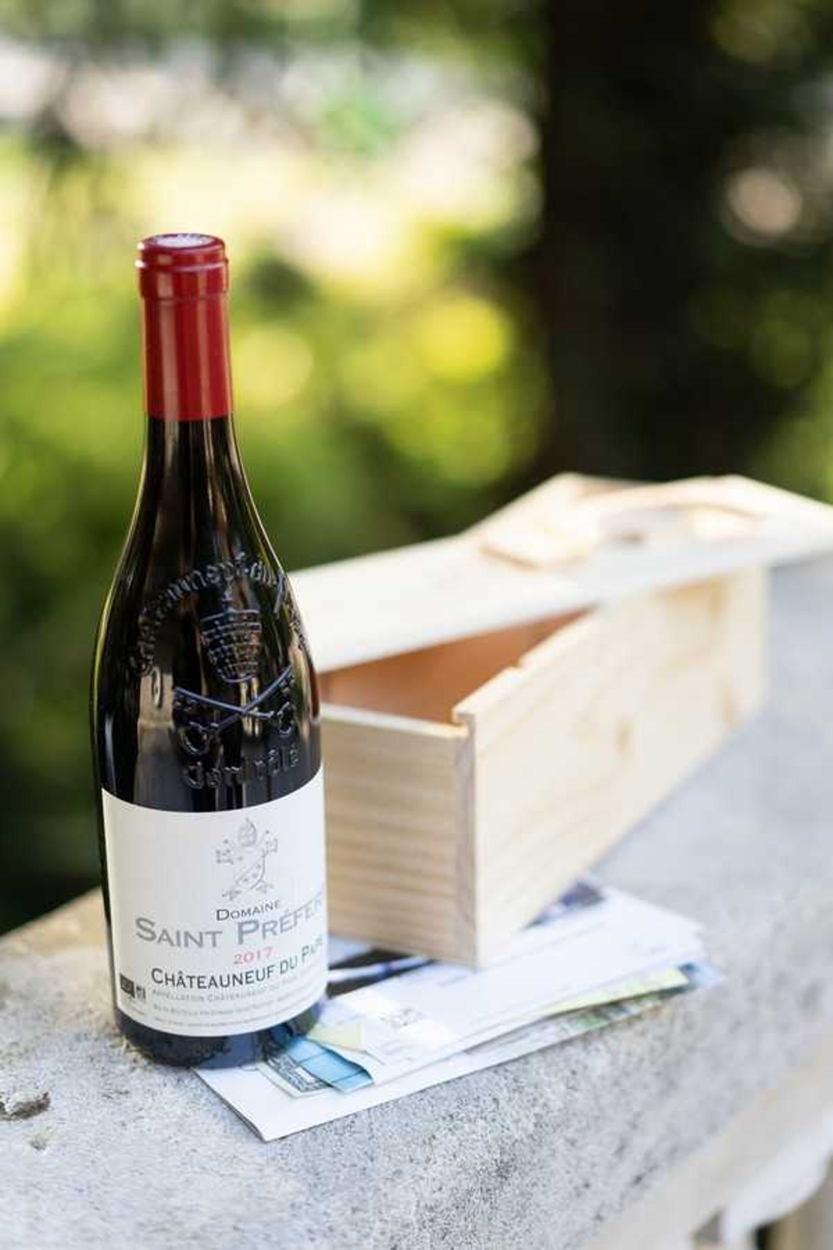 A close-up detail shot of a bottle of Domaine Saint Prefert 2017 Châteauneuf-du-Pape red wine with a red foil cap, placed on a stone ledge alongside an open light wood single-bottle gift box and what appears to be printed cards or paperwork beneath them. The background is a blurred outdoor garden setting with green foliage. This appears to be a wedding gift or favor detail, likely styled as part of a wine country or vineyard-themed wedding. The composition is a near-flat product-style portrait with shallow depth of field.