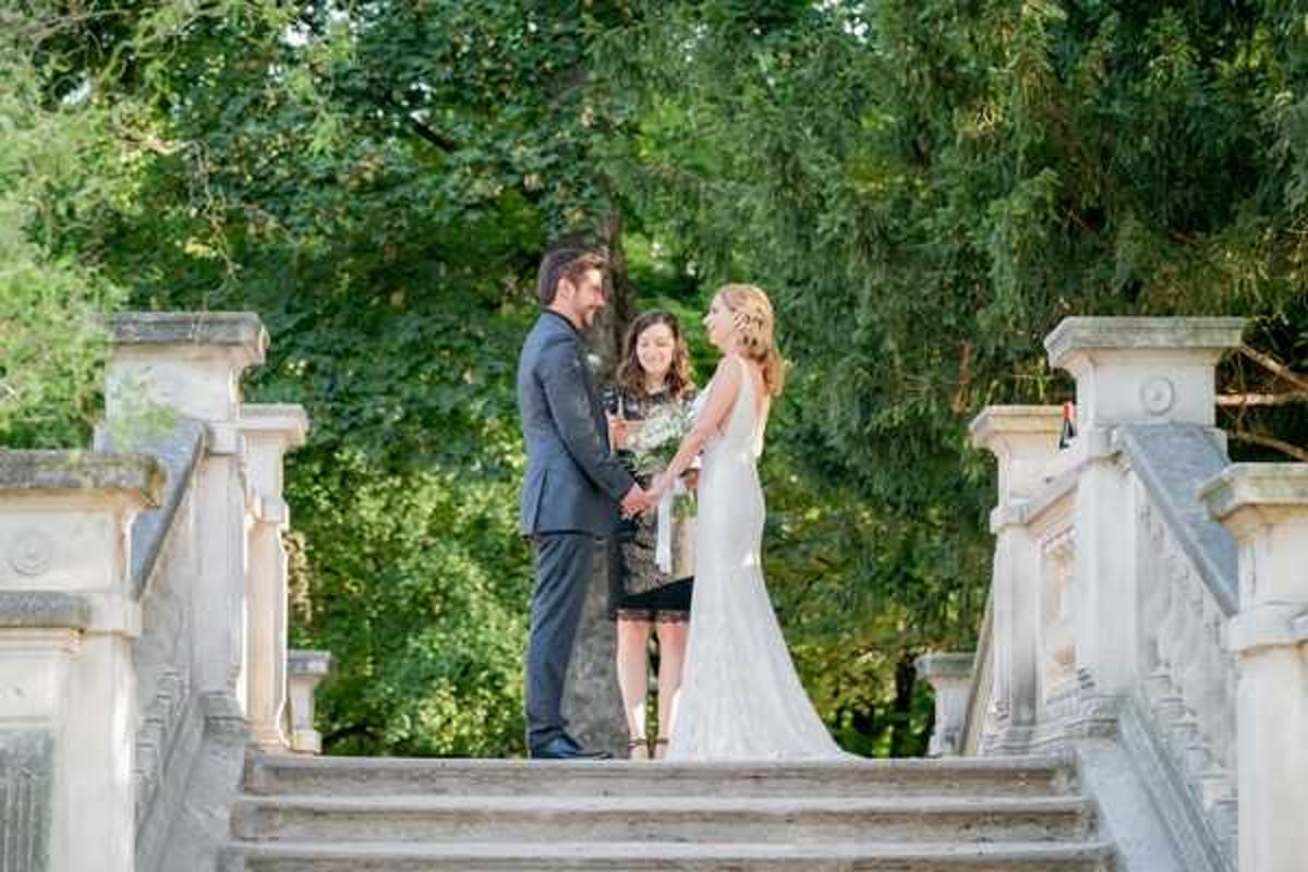 An outdoor wedding ceremony taking place on a wide stone staircase with ornate balustrades, likely at a French chateau or formal estate garden. The bride wears a fitted, backless white lace gown and holds a small bouquet with white blooms and greenery, while the groom is dressed in a dark navy suit; they face each other holding hands. A female officiant in a dark patterned dress stands between them reading from a booklet. Wide shot framing the couple and officiant centrally on the stairs with mature trees forming a dense green backdrop.