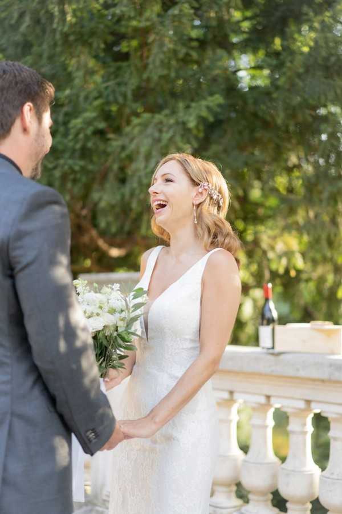 A bride and groom share a candid moment on an outdoor stone balustrade terrace, with the bride laughing openly while holding the groom's hands. The bride wears a fitted sleeveless white V-neck dress with a textured fabric, carries a small bouquet of white flowers and greenery, and has her hair styled in soft waves with a floral hair accessory. The groom, seen from behind, wears a charcoal grey suit. In the background, a wine bottle and a wooden box are visible on the stone balustrade, suggesting a wine box ceremony element. The shot is a mid-length portrait with soft natural lighting.