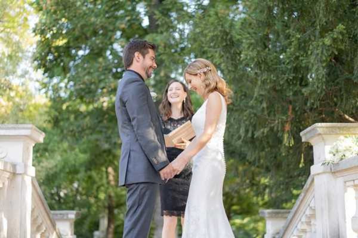 An outdoor wedding ceremony captured in a medium portrait shot, showing a couple exchanging vows on what appears to be a stone balustrade or terrace, likely at a French chateau or formal garden. The groom wears a charcoal grey suit with a white boutonniere, and the bride wears a fitted sleeveless white lace gown with a delicate floral headpiece, her blonde hair styled in a loose updo. A female officiant in a black lace dress stands between them holding a gold-covered book, smiling warmly as the couple hold hands and laugh together. White floral arrangements are partially visible on the stone balustrade flanking the trio.