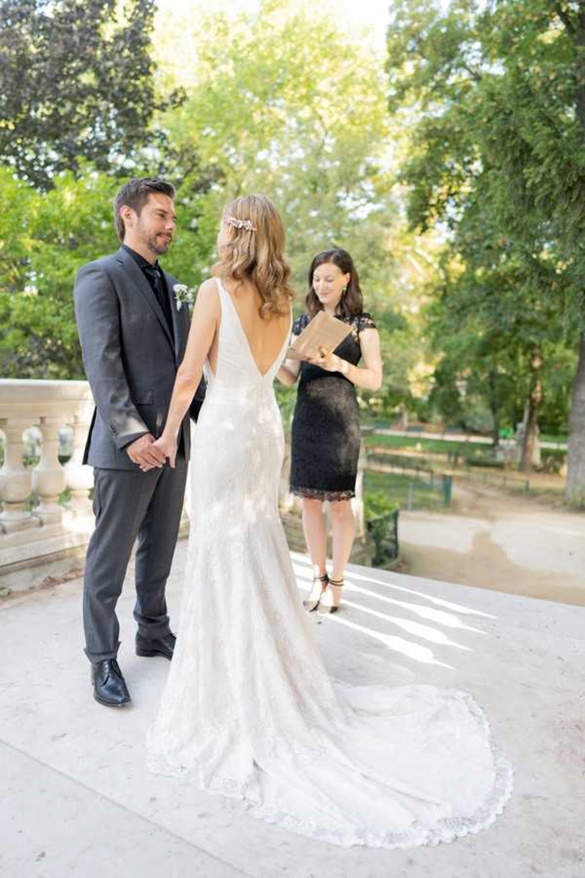 An outdoor wedding ceremony taking place on a stone terrace with a classical balustrade railing, surrounded by mature trees. The couple stands facing each other holding hands — the groom wears a charcoal grey suit with a dark tie and a white boutonniere, while the bride wears a fitted ivory lace gown with a deep V-back, a long train, and a floral hair accessory. A female officiant dressed in a black lace knee-length dress stands opposite them, holding an open tan/gold book and reading from it. The setting and architecture suggest a formal garden or park environment, consistent with a classic French venue. Portrait-style shot taken from behind the bride, showing the full length of her gown's train.