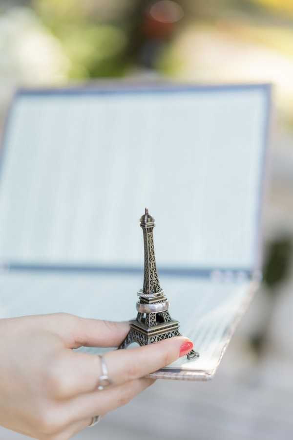 A close-up detail shot of a hand with red-painted nails and a silver ring holding a small bronze-toned Eiffel Tower miniature figurine, which also holds a wedding band around its base. The figurine appears to be used as a wedding ring holder or decorative favor. In the soft-focus background, a white and blue-bordered card or menu is visible, suggesting a table setting at an outdoor reception. The Paris-themed styling detail indicates a French or Paris-inspired wedding aesthetic.