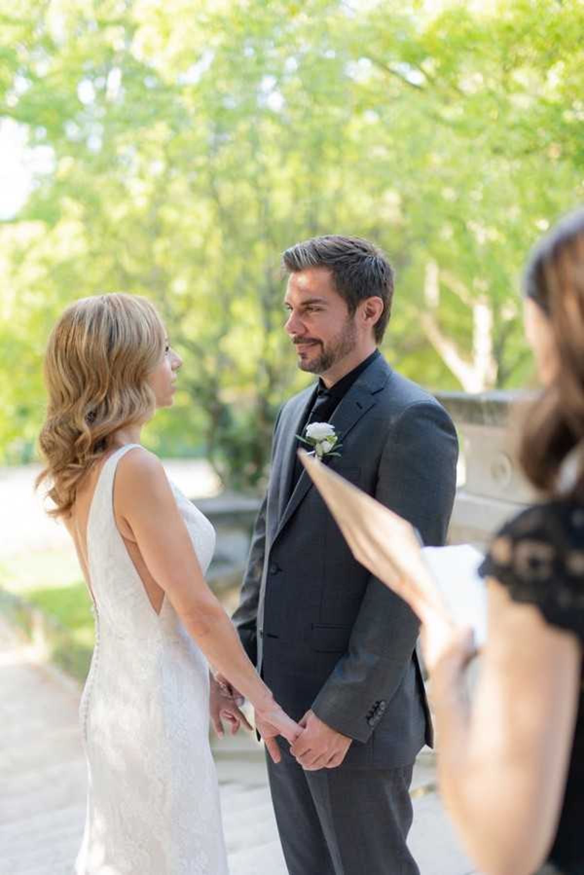 An outdoor wedding ceremony showing a couple exchanging vows while holding hands and facing each other. The bride wears a fitted sleeveless white lace gown with an open back, and the groom wears a charcoal grey suit with a black shirt and tie, accented by a white rose boutonniere. In the foreground, partially blurred, is an officiant in a black lace top holding papers. The setting is a paved outdoor area with trees in the background, suggesting a garden or courtyard venue. The shot is a medium portrait taken from behind and to the side of the bride, with natural daytime lighting.