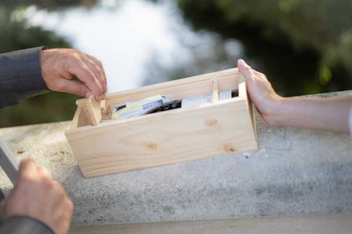 Close-up detail shot of a couple's hands handling an open natural pine wood box placed on a stone surface during what appears to be an outdoor ceremony ritual. The box contains small items including what appear to be letters or notes and other personal objects, suggesting a unity ceremony wine box or time capsule tradition. One hand, likely the groom's, is dressed in a grey suit sleeve, while the other hand, likely the bride's, rests on the side of the box. The background is softly blurred with greenery and a hint of water.