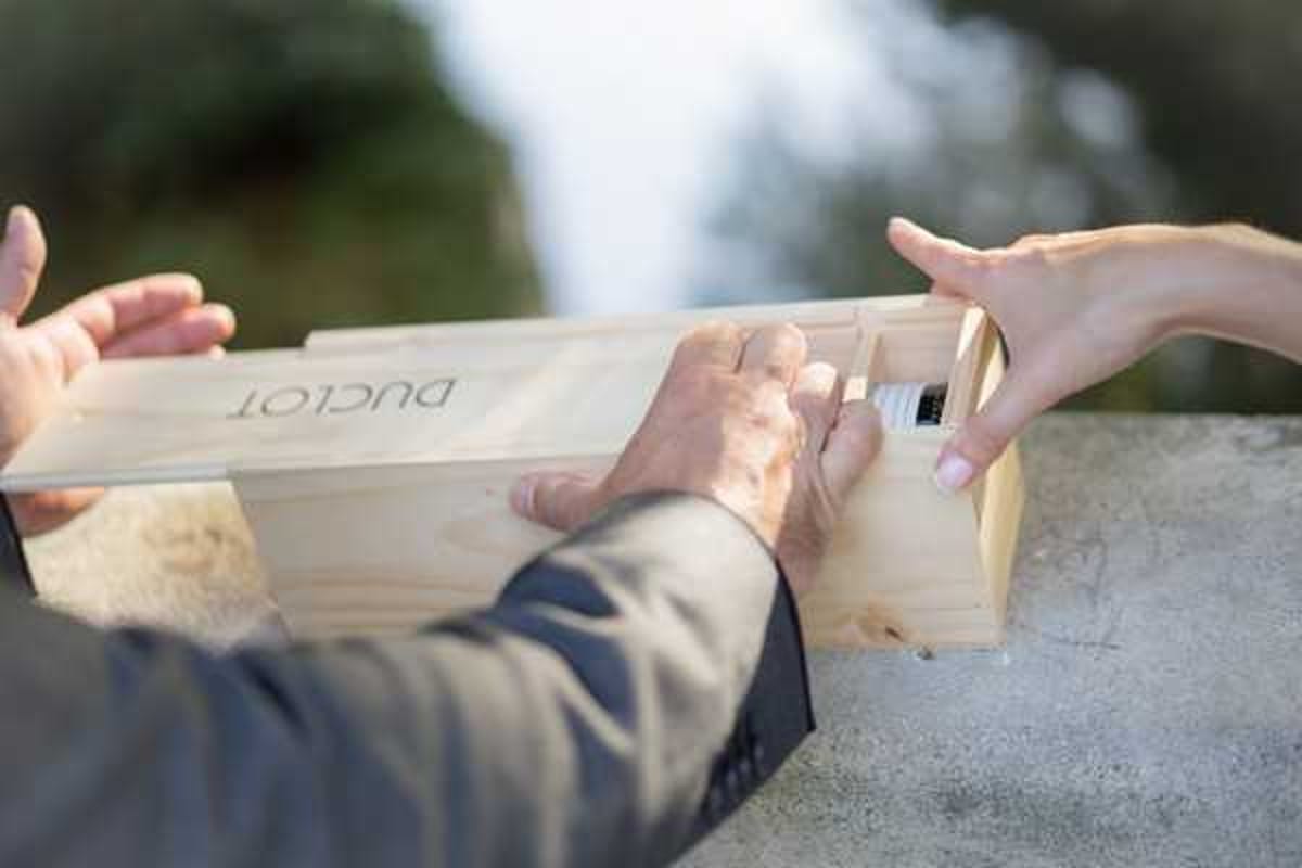 A close-up detail shot of two people — likely the couple — handling a wooden wine box branded with the name 'Duclot,' a Bordeaux wine merchant. One person, wearing a dark suit jacket, holds the box from one side while a second person with manicured nails reaches in from the other side, appearing to place or retrieve a small item, possibly a letter or capsule as part of a wine box unity ceremony. The setting appears to be outdoors on a stone or concrete surface, with a soft blurred background. The composition focuses tightly on the hands and the wooden box, emphasizing this as a symbolic ritual moment during the wedding.