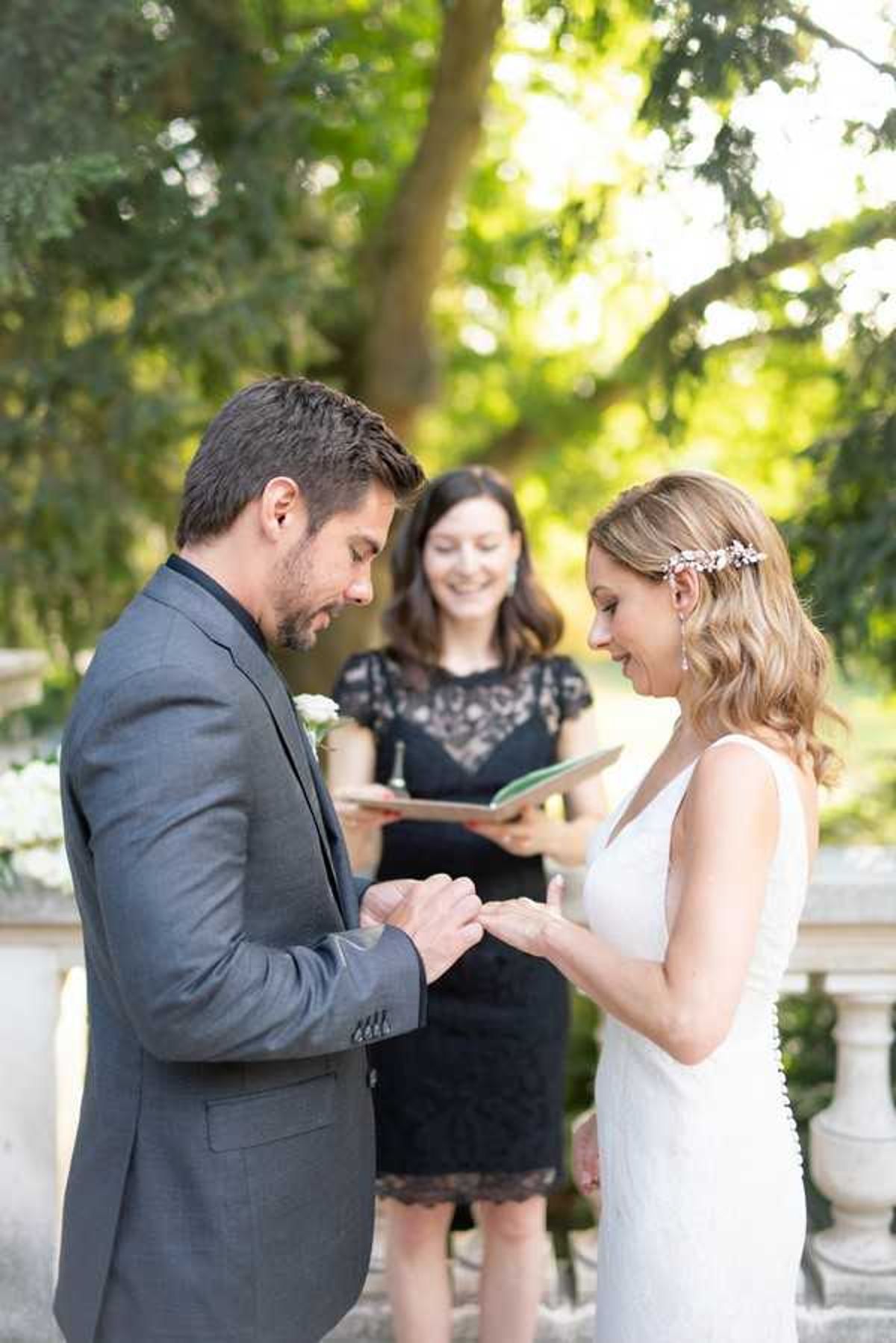 The groom is placing a ring on the bride's finger during an outdoor ceremony ring exchange, with a smiling officiant standing behind them holding an open book. The setting appears to be a garden or estate grounds, with stone balustrade details visible in the background. The groom wears a charcoal grey suit with a white boutonniere and dark tie, while the bride wears a sleeveless white V-neck fitted gown and a delicate floral hair accessory with drop earrings. The officiant is dressed in a black lace short-sleeve dress. The shot is a medium portrait framing all three figures, with the couple in the foreground in sharp focus and the background softly blurred.