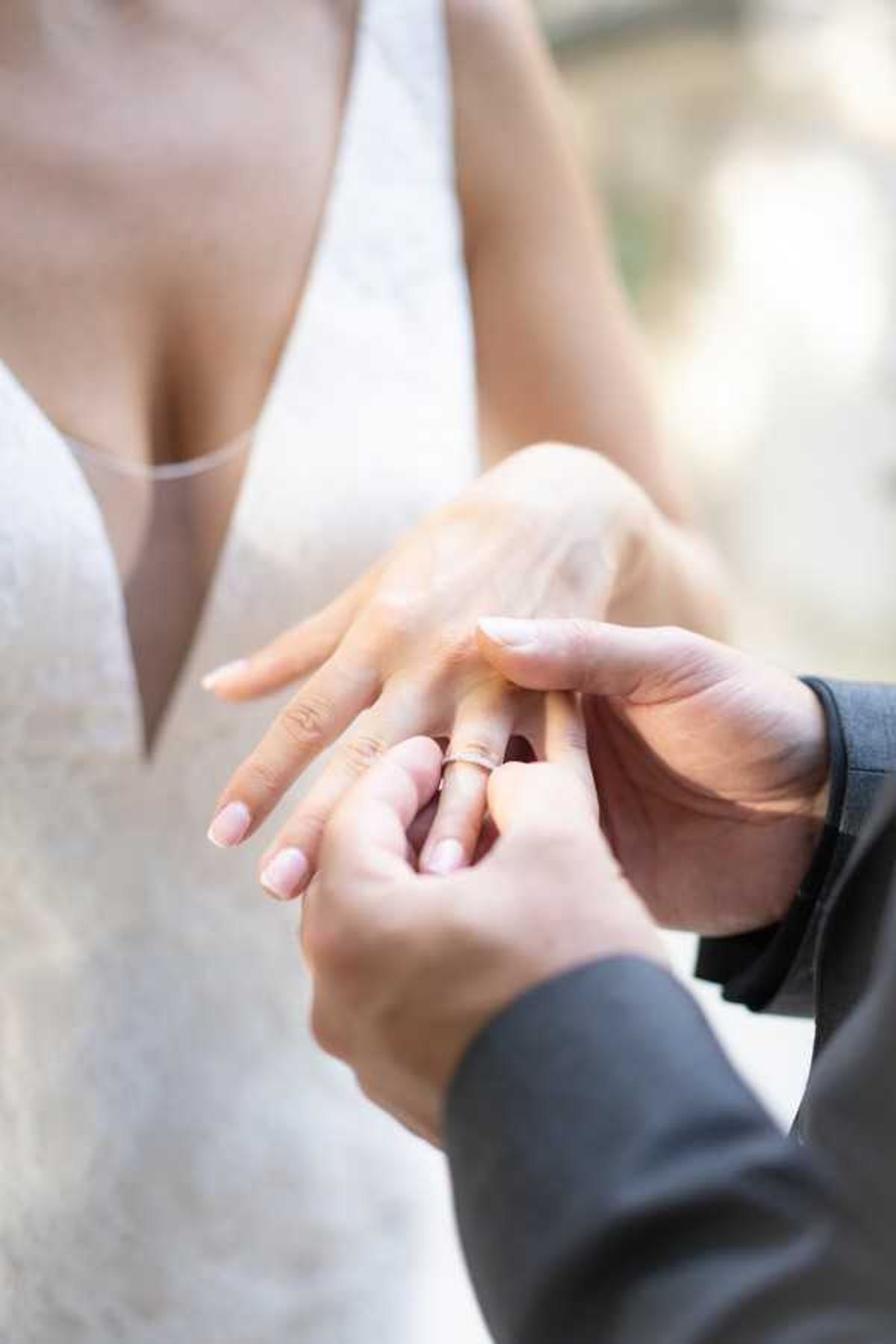 Close-up detail shot of the ring exchange moment during a wedding ceremony, showing the groom placing a thin gold band onto the bride's finger. The bride is wearing a white lace gown with a deep V-neckline, and the groom is dressed in a dark grey suit. The background is fully blurred, keeping the focus entirely on the hands and ring. The composition is a tight portrait-style detail shot with soft natural lighting.