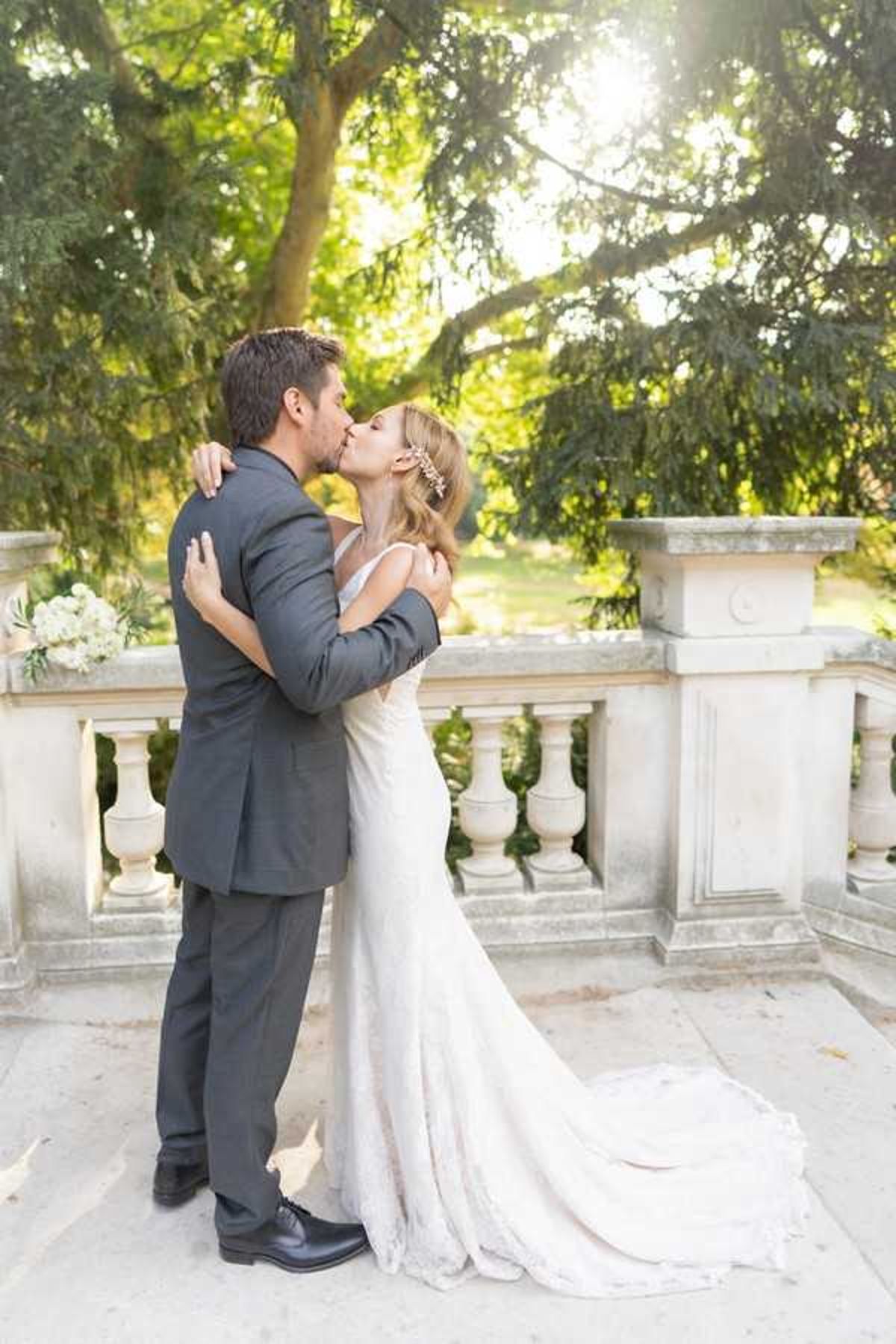 A couple shares a kiss during an outdoor portrait session on a stone balustrade terrace, likely at a chateau or formal garden. The bride wears a fitted ivory lace gown with a short train and a delicate floral hair accessory, while the groom is dressed in a charcoal grey suit with black dress shoes. A small bouquet of white blooms, possibly peonies or roses with greenery, is visible resting on the stone railing to the left. The shot is a full-length portrait with soft backlit natural light filtering through the trees, giving the image a warm, golden tone.