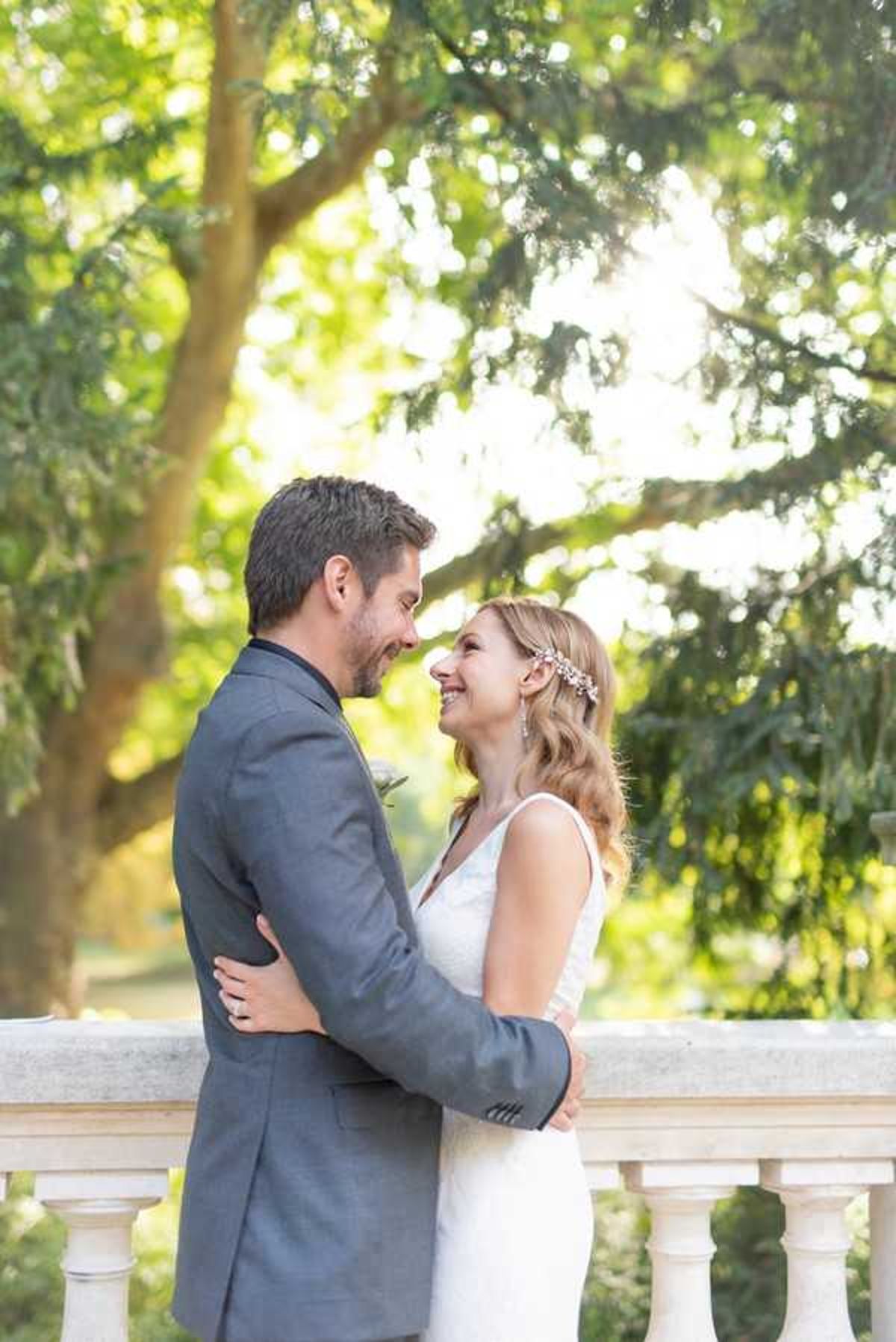 A couple portrait taken outdoors on a terrace or balustrade featuring white stone baluster railings. The bride wears a sleeveless white fitted gown and a delicate floral hair accessory with small pink and crystal blooms, her blonde hair styled in loose waves. The groom wears a charcoal grey suit with a dark lapel. The two are embracing and facing each other closely, both smiling, in a relaxed and candid pose. The shot is a medium portrait framed with soft bokeh from the surrounding trees, with warm golden backlight filtering through the foliage.