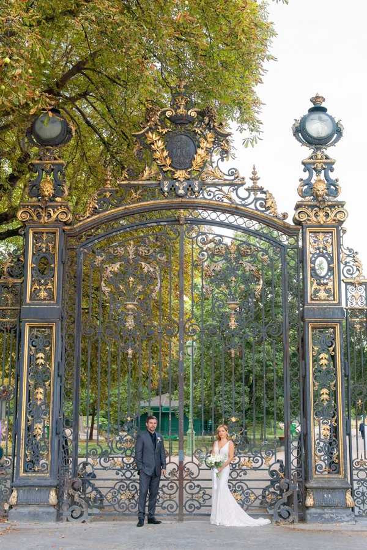 A bride and groom stand side by side in front of large ornate wrought-iron gates with gold-gilt detailing, scrollwork, and a crest at the top — consistent with the gates of Parc Monceau in Paris. The groom wears a charcoal grey suit with a dark tie and a white boutonniere. The bride wears a fitted, slim-cut ivory gown with a V-neckline and holds a white and green bouquet, likely white roses or peonies with greenery. The portrait is taken as a medium-wide shot, positioning the couple small relative to the grand scale of the gates, emphasizing the ornate architecture. The overall styling is classic and formal.