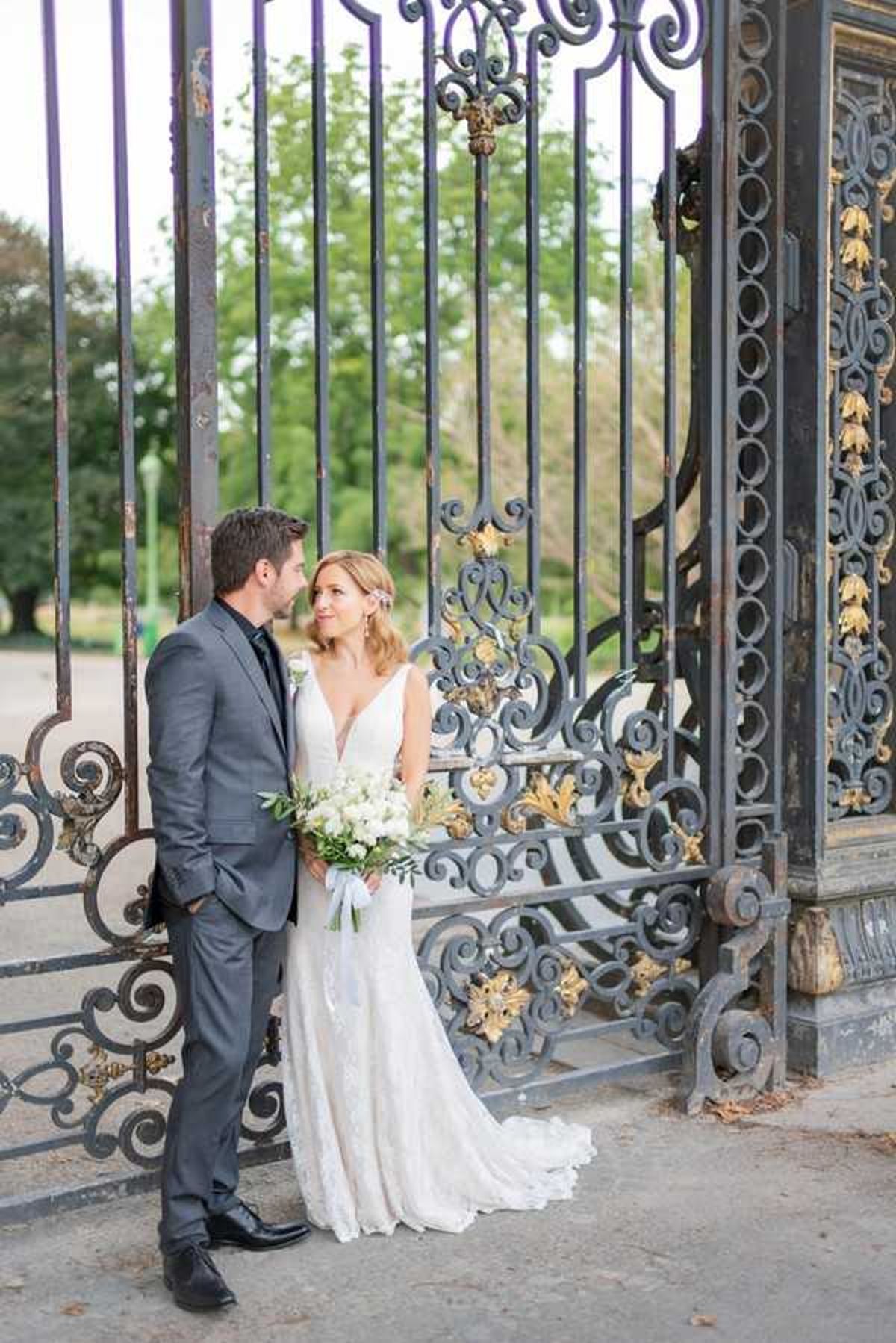 A couple portrait taken outdoors in front of large ornate wrought-iron gates featuring gold-painted decorative leaf and scroll detailing. The bride wears a fitted, deep V-neck lace gown with a short train and holds a bouquet of white flowers — including what appear to be white tulips or ranunculus — with greenery and a light blue ribbon wrap. The groom wears a charcoal grey suit with a dark tie and stands close to the bride, leaning in toward her while she looks up at him. The styling is classic and refined, with a neutral white-and-green floral palette. Medium portrait shot, both figures visible full-length.