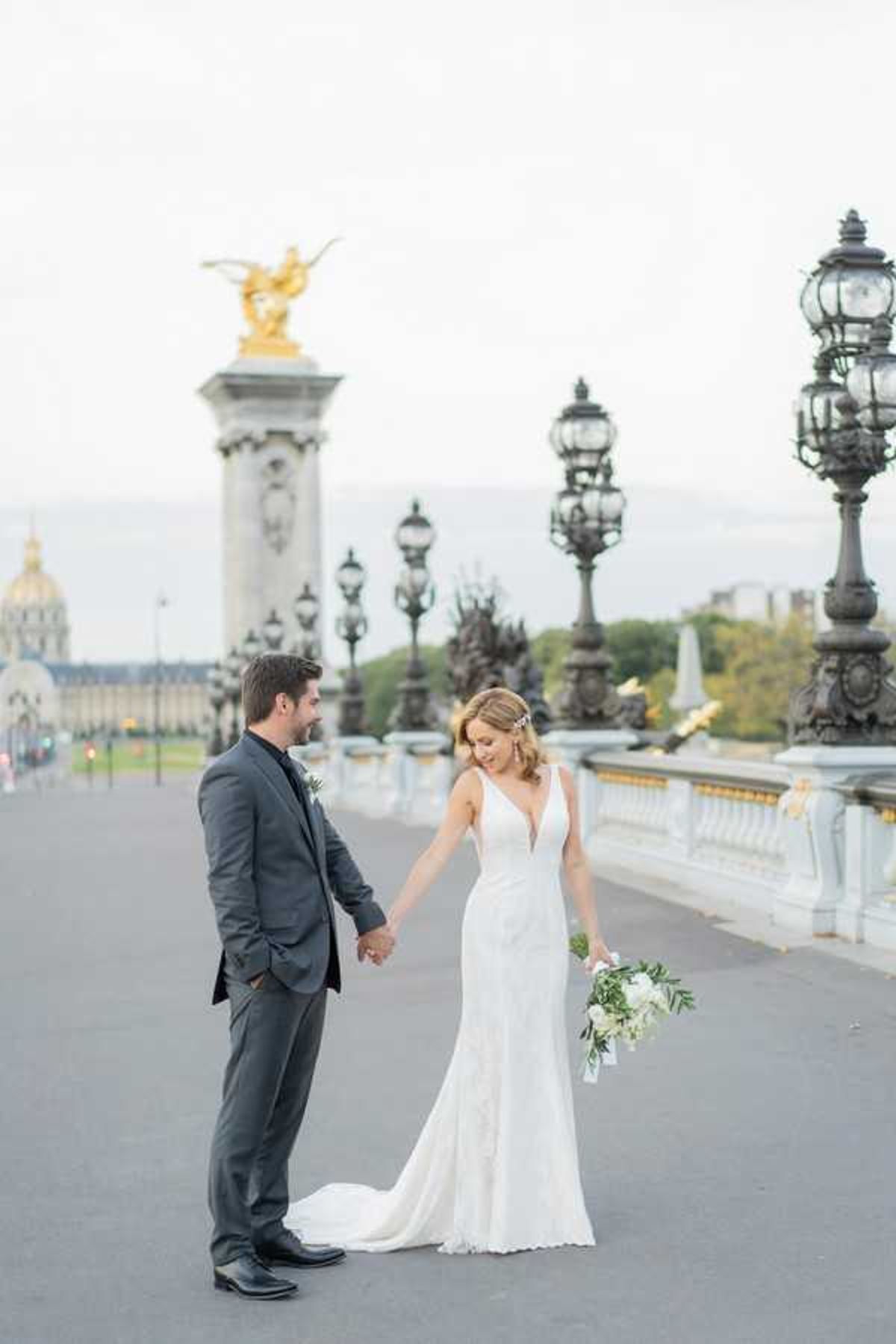 A bride and groom portrait taken on the Pont Alexandre III bridge in Paris, with the gilded sculpture columns and ornate black iron lamp posts of the bridge visible in the background, along with the Hôtel des Invalides dome to the left. The groom wears a charcoal grey suit with a white boutonniere, while the bride wears a fitted, deep V-neck white gown with a lace hemline and short train. She carries a loose bouquet of white blooms and greenery, including what appears to be white roses and eucalyptus. The couple holds hands facing each other, with the bride glancing down and smiling. The shot is a full-length portrait taken at ground level during soft, overcast late-day light.
