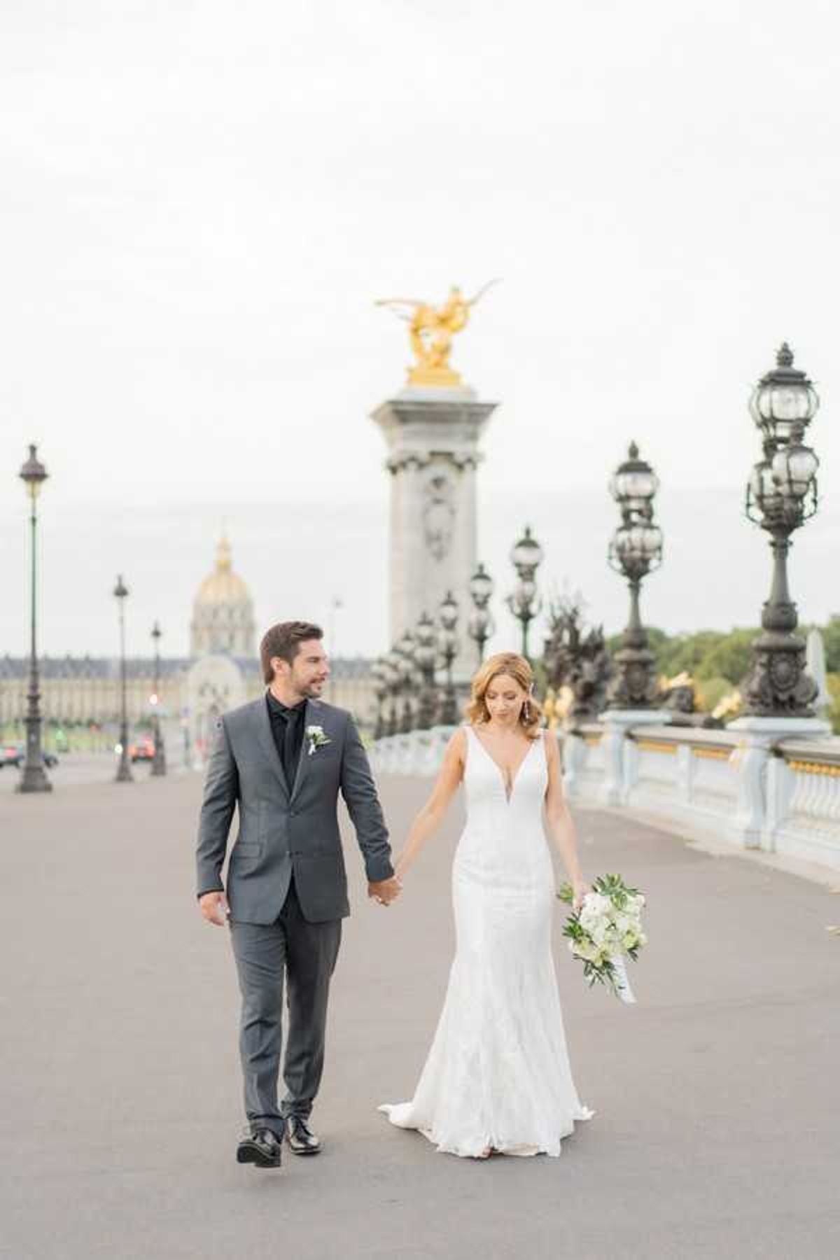 A couple portrait taken outdoors on the Pont Alexandre III bridge in Paris, with the gilded bronze sculpture atop one of the bridge's ornate stone pillars and the dome of Les Invalides visible in the background. The groom wears a charcoal grey suit with a black dress shirt and a white floral boutonniere, while the bride wears a fitted, deep V-neck lace gown with a slight fishtail hem. She carries a loose bouquet of white flowers and greenery, looking downward as the groom looks toward her with a smile. The shot is a medium-wide walking portrait with soft, overcast natural light.
