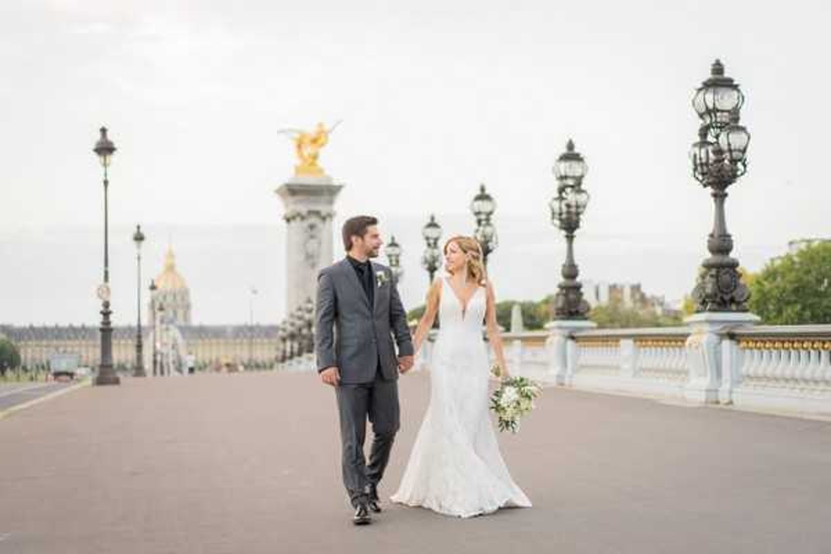 A couple portrait taken outdoors on the Pont Alexandre III bridge in Paris, with the gilded statue columns and ornate iron lampposts lining the bridge visible in the background, and Les Invalides dome faintly visible in the distance. The bride wears a fitted, V-neck lace wedding gown with a slight train and holds a loose bouquet of white and green florals with eucalyptus. The groom wears a charcoal grey suit with a black tie. The two are walking hand-in-hand and looking at each other, shot as a mid-length portrait with the bridge extending into the background.