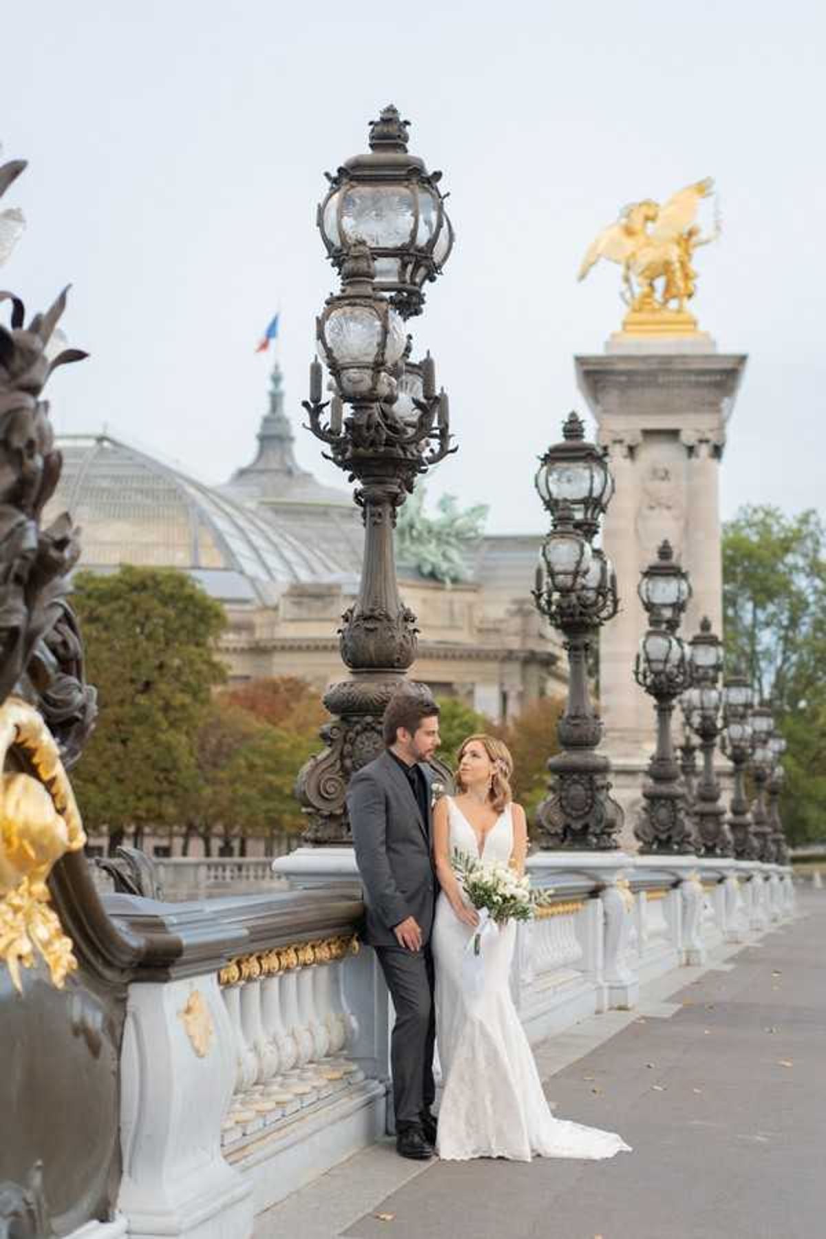 A couple poses for a wedding portrait on the Pont Alexandre III bridge in Paris, with the Grand Palais visible in the background. The bride wears a fitted white lace gown with a deep V-neckline and holds a bouquet of white flowers with greenery and eucalyptus, while the groom is dressed in a dark charcoal suit with a black shirt. The ornate cast-iron lampposts and gold-gilded sculptures characteristic of the bridge's Belle Époque design frame the couple, with a gilded winged figure atop one of the bridge's stone pillars visible to the right. The shot is a full-length portrait taken from a slight distance, capturing both the couple and the architectural details of the bridge.