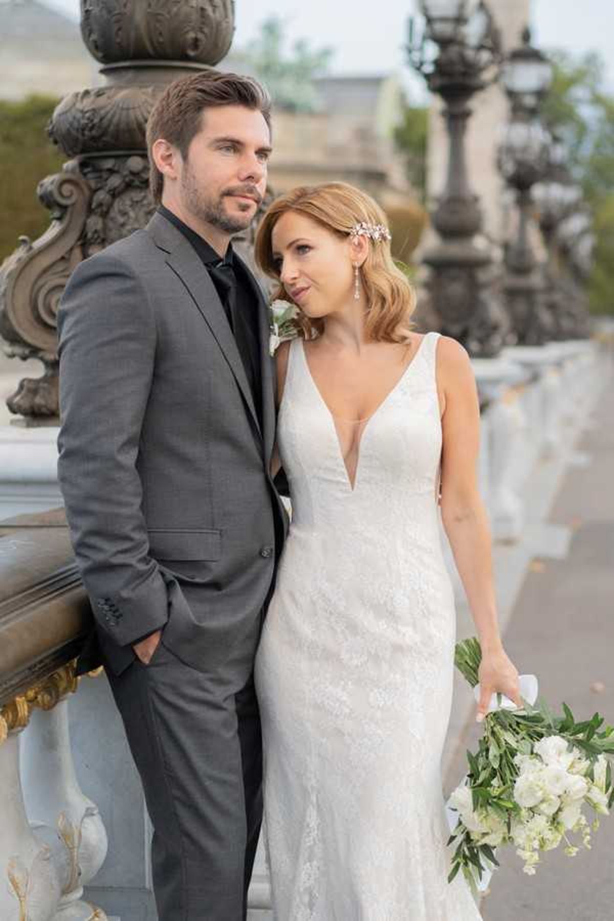 A couple portrait taken outdoors on what appears to be the Pont Alexandre III bridge in Paris, identifiable by the ornate gilded lampposts and carved stone balustrade in the background. The groom wears a charcoal grey suit with a black dress shirt and black tie, while the bride wears a fitted lace gown with a deep V-neckline; she holds a loose bouquet of white flowers with green foliage and wears a small floral hair accessory. The bride looks up at the groom while he gazes off to the side. This is a mid-length portrait shot with a shallow depth of field that blurs the Parisian architectural details behind them.