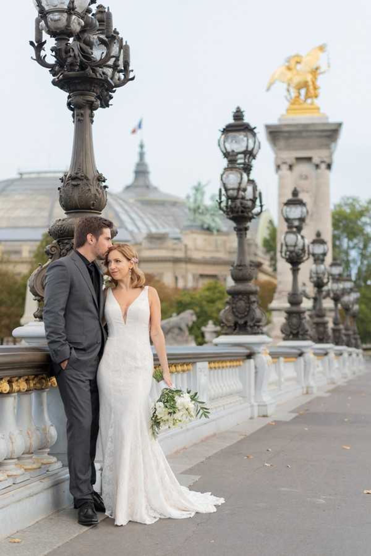A couple portrait taken outdoors on the Pont Alexandre III bridge in Paris, with the Grand Palais visible in the background along with the bridge's iconic ornate bronze lamp posts and a gilded sculpture atop one of the stone pillars. The groom, wearing a charcoal grey suit with a dark tie, leans against the bridge railing and kisses the bride on the temple. The bride wears a fitted lace gown with a deep V-neckline and a short train, and holds a loose bouquet of white flowers and greenery including what appear to be white peonies or ranunculus with eucalyptus. The composition is a full-length couple portrait with the bridge's architecture providing strong leading lines into the background.