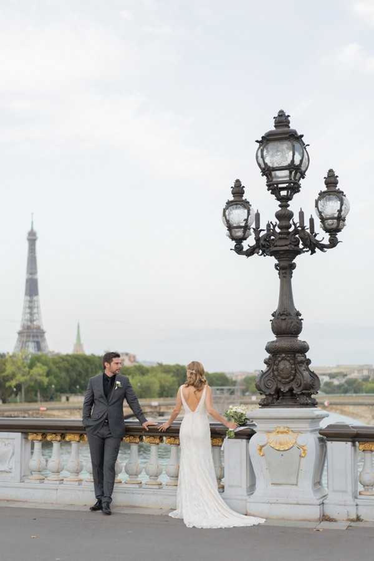 A couple poses for wedding portraits on the Pont Alexandre III bridge in Paris, with the Eiffel Tower visible in the background. The groom wears a charcoal grey suit with a black tie and white boutonniere, while the bride wears a form-fitting ivory gown with a deep V-back and a short train, holding a small white bouquet. The ornate bronze and gold lamp post of the bridge is prominently featured in the foreground right, with the bridge's white stone balustrade and gold shell detailing visible. Wide portrait shot taken at a slight distance, capturing both figures full-length with the Paris skyline as backdrop.