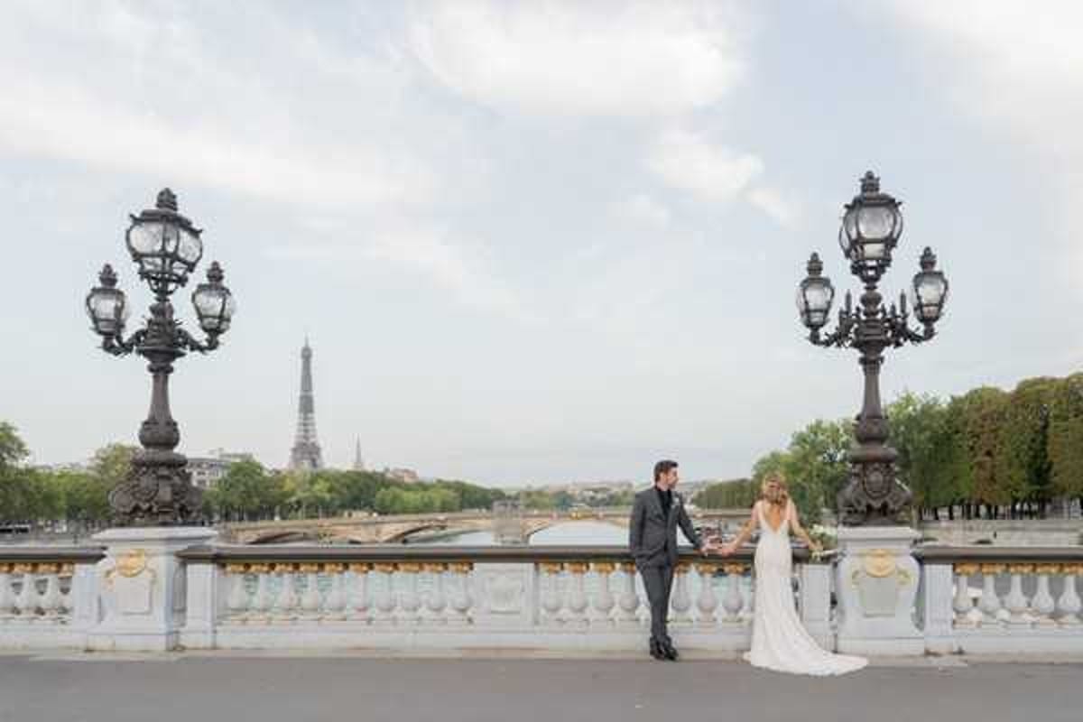 A bride and groom pose together on the Pont Alexandre III bridge in Paris, with the Eiffel Tower visible in the background along the Seine River. The groom wears a dark charcoal suit and the bride wears a fitted, low-back white lace gown, holding a small bouquet with warm-toned florals. The couple stands between two ornate cast-iron lamp posts characteristic of the bridge's Belle Époque architecture, facing each other in a relaxed portrait pose. This is a wide shot taken during daytime with soft, even natural light, capturing both the couple and the Parisian cityscape.