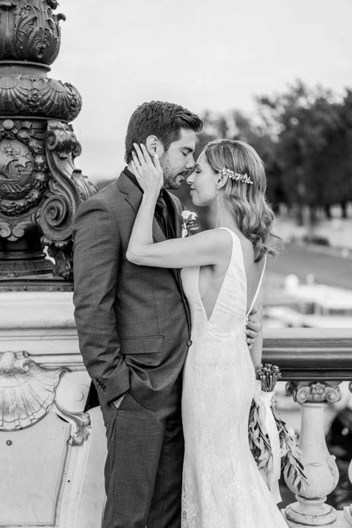 A black-and-white couple portrait taken outdoors on an ornate stone balustrade or terrace, likely at a French chateau or grand civic building, with heavily carved baroque-style architectural details visible in the foreground. The bride wears a low-back lace gown with thin straps and a deep V-back, her wavy hair styled down and adorned with a delicate floral or leaf hair accessory; she holds the groom's face gently with one hand as they lean toward each other, foreheads nearly touching. The groom wears a fitted suit with a boutonniere on his lapel, hands relaxed at his sides. The image is a medium close-up portrait with soft contrast and light tones, with a blurred background suggesting an elevated outdoor setting with distant trees and grounds visible. Potential venue feature image.