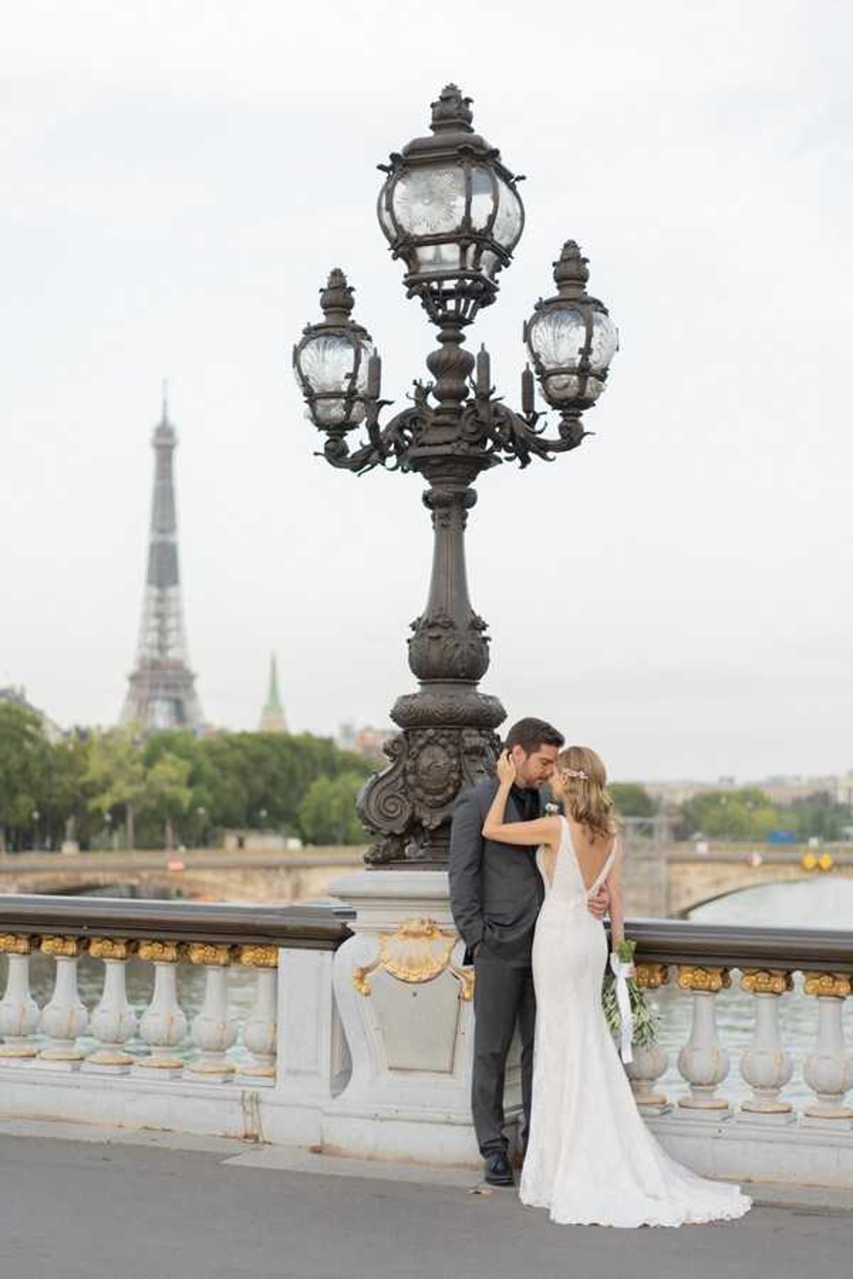 A couple portrait taken outdoors on the Pont Alexandre III bridge in Paris, with the Eiffel Tower visible in the soft-focus background. The bride wears a fitted ivory gown with a low open back and a short train, her hair accessorized with a floral crown, and she holds a loose greenery-forward bouquet. The groom is dressed in a charcoal grey suit with a dark tie. The two stand close together, foreheads nearly touching, positioned beside one of the bridge's ornate bronze multi-lantern lampposts with gold-detailed stone balustrade in the foreground. The shot is a full-length portrait taken from behind and slightly to the side, capturing the architectural details of the bridge alongside the Seine River and city skyline.