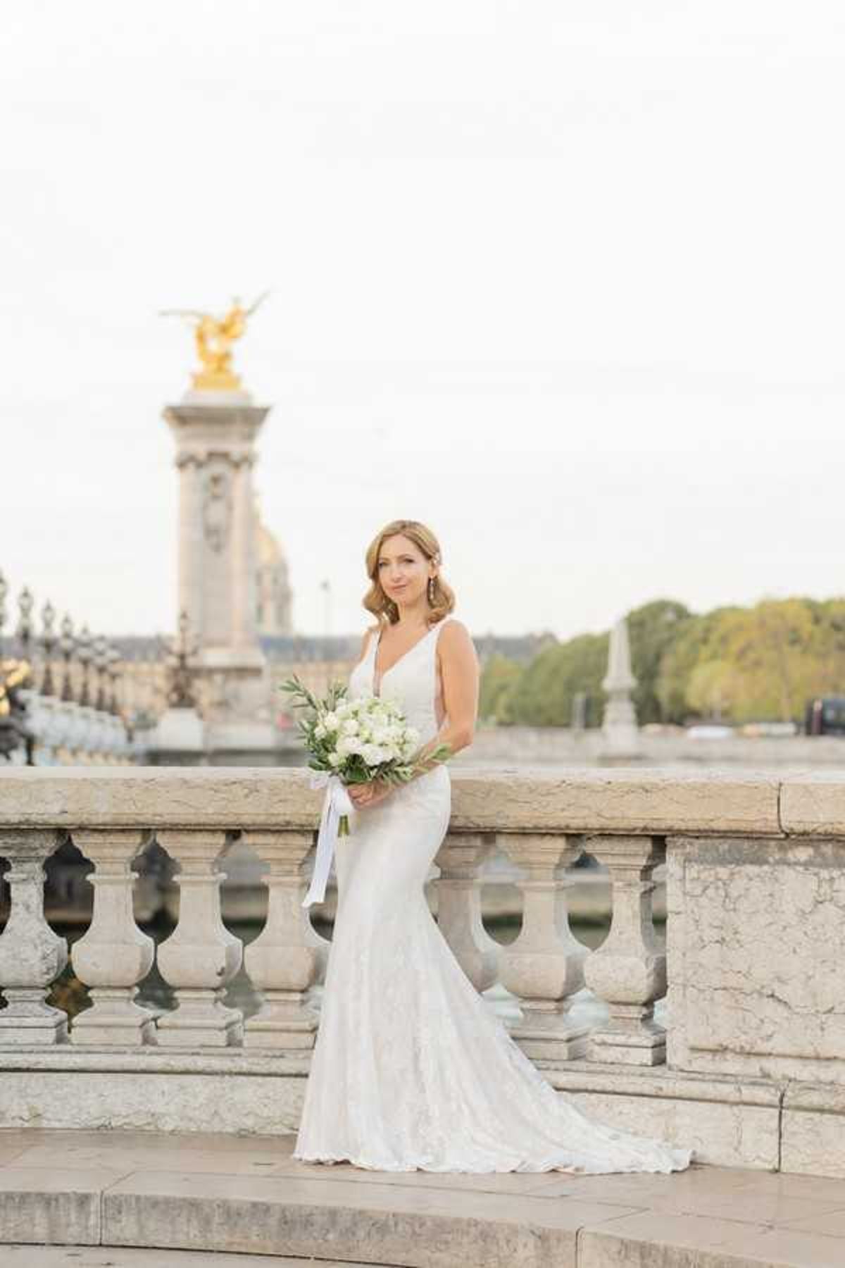 A bridal portrait taken outdoors on the Pont Alexandre III in Paris, with the bridge's ornate stone balustrade and a gilded bronze sculpture atop one of its iconic columns visible in the background. The bride stands alone, facing the camera, wearing a fitted ivory lace gown with a V-neckline, sleeveless silhouette, and a slight train. She holds a bouquet of white ranunculus and roses with green eucalyptus and olive foliage, tied with a white satin ribbon. The image is a full-length portrait shot taken in soft, diffused natural light with a shallow depth of field that keeps the Parisian architecture gently out of focus behind her.