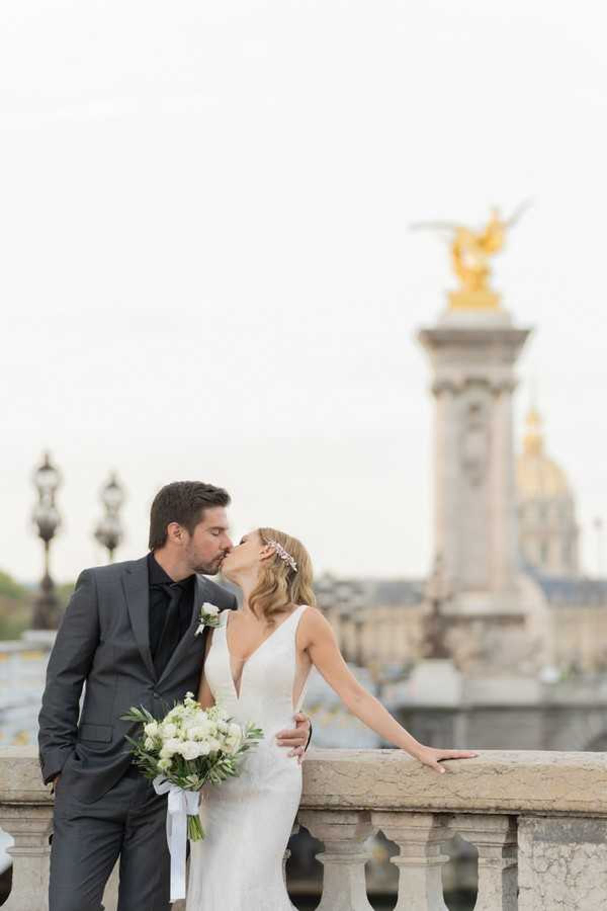 A couple shares a kiss during outdoor wedding portraits on the Pont Alexandre III bridge in Paris, with one of the bridge's iconic gilded bronze sculptures and the dome of Les Invalides visible in the soft-focus background. The bride wears a form-fitting white gown with a deep V-neckline and a delicate floral hair piece, and carries a bouquet of white ranunculus and garden roses with green foliage tied with a white ribbon. The groom wears a charcoal grey suit with a black dress shirt and black tie, with a small white boutonnière on his lapel. The image is a mid-distance portrait with a shallow depth of field that keeps the couple sharp against the blurred Parisian architectural backdrop.