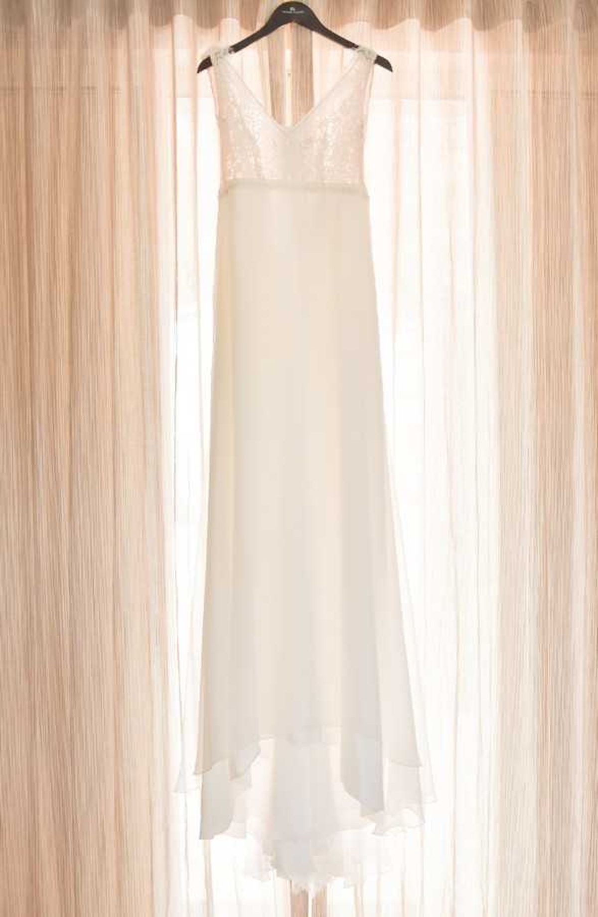 A getting-ready detail shot of a white wedding dress hanging on a black hanger against sheer, warm-toned curtains backlit by natural light. The gown features a V-neckline bodice with sequin or lace embellishment on the upper portion, transitioning to a smooth, flowing chiffon or crepe skirt with a subtle layered hem. The dress has a sleeveless, minimalist silhouette with a slightly asymmetric handkerchief-style hemline. The composition is a full-length vertical shot centered on the dress.