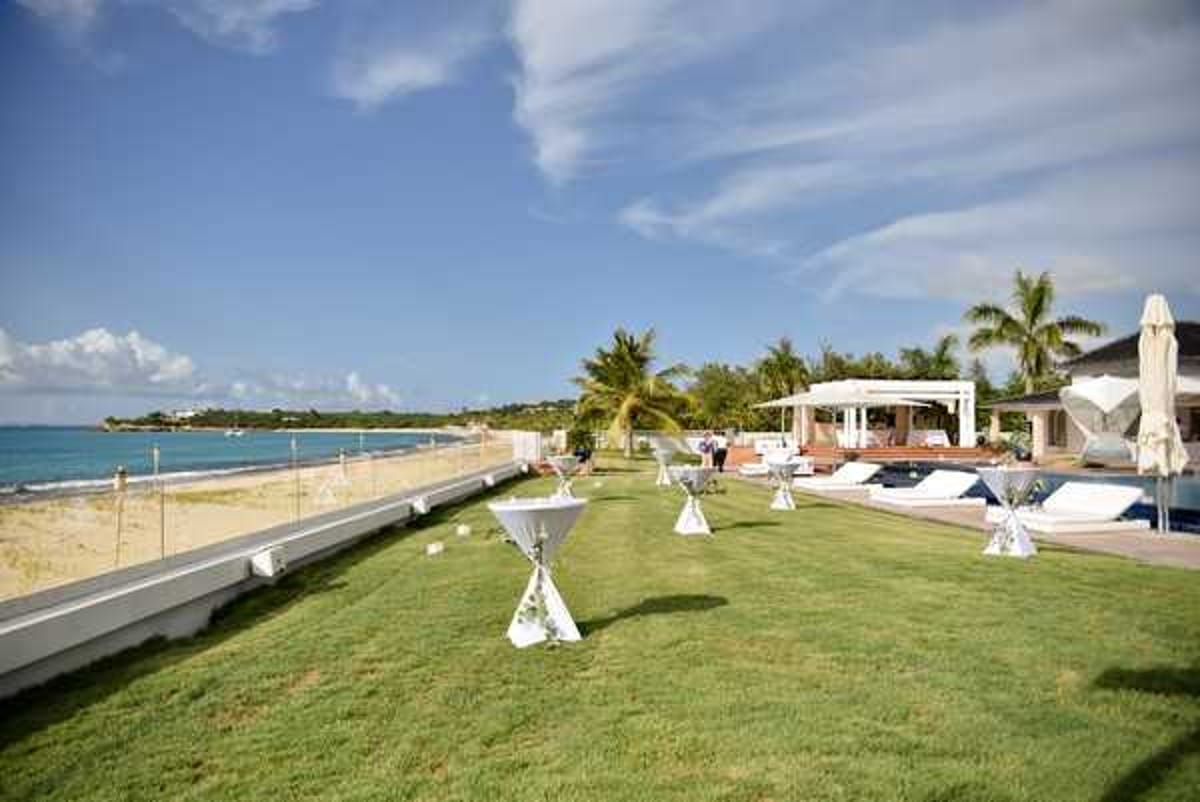 A cocktail hour or pre-reception setup is shown at an outdoor beachfront venue, with several white spandex-covered cocktail tables with small floral centerpieces arranged across a manicured lawn. The setting features a pool area with white sun loungers, a white pergola structure, and closed white market umbrellas in the background, alongside a sandy beach and turquoise water. A few guests or staff members are visible in the mid-ground near the pool area. The overall decor palette is white and minimal, consistent with a clean, modern tropical wedding aesthetic. Wide establishing shot. Potential venue feature image.