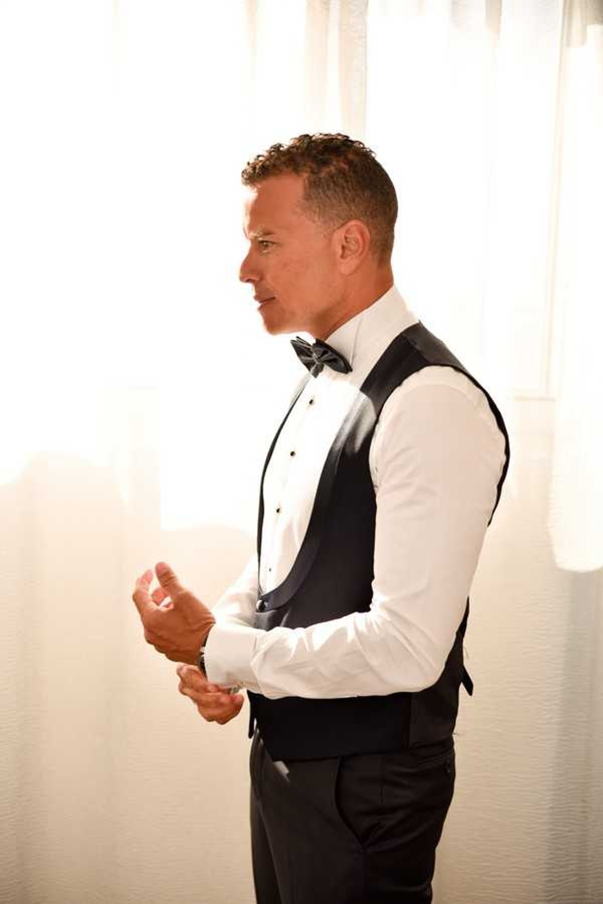 A groom getting ready, photographed in profile as he fastens his cufflinks. He is dressed in a classic black tuxedo waistcoat with satin lapels, a white formal dress shirt, and a black bow tie, paired with black trousers. The setting is an indoor room with sheer white curtains backlit by bright natural light, creating a warm, overexposed background. The shot is a mid-length portrait with a clean, minimal composition.