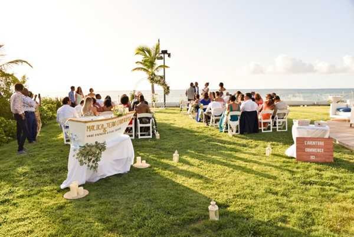 An outdoor wedding ceremony taking place on a grass lawn directly overlooking the ocean, shot as a wide angle from behind and to the side of the seating area. Approximately 30-40 guests are seated in white folding chairs arranged in rows, facing the officiant and couple at the front. In the foreground, a welcome or signing table is draped in a white cloth with a personalized banner reading 'Malika & Jean Christophe' and decorated with a greenery arrangement; white lanterns are placed on the grass nearby. A wooden sign reading 'L'Aventure Commence' is positioned to the right. The decor style is relaxed and tropical, with minimal florals and a natural color palette. The ceremony is set at golden hour, with warm low sunlight and a palm tree visible in the background against the sea horizon.