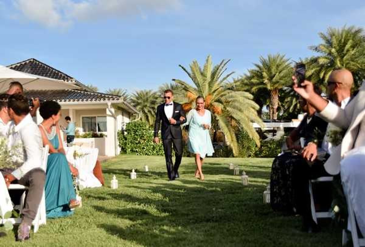 A groom in a black tuxedo with a white dress shirt and black bow tie walks down the outdoor ceremony aisle escorted by a woman in a knee-length light blue dress, likely his mother. The ceremony is set on a manicured lawn in front of a low white villa-style building with a dark tiled roof, flanked by tall palm trees. White folding chairs are arranged on either side of the grass aisle, lined with small white lanterns as aisle markers, with approximately 15–20 guests visible; a bridesmaid in a teal floor-length dress is seated on the left. The shot is a medium wide angle taken from ground level, capturing the processional moment with guests watching and one guest on the right holding up a phone to record.