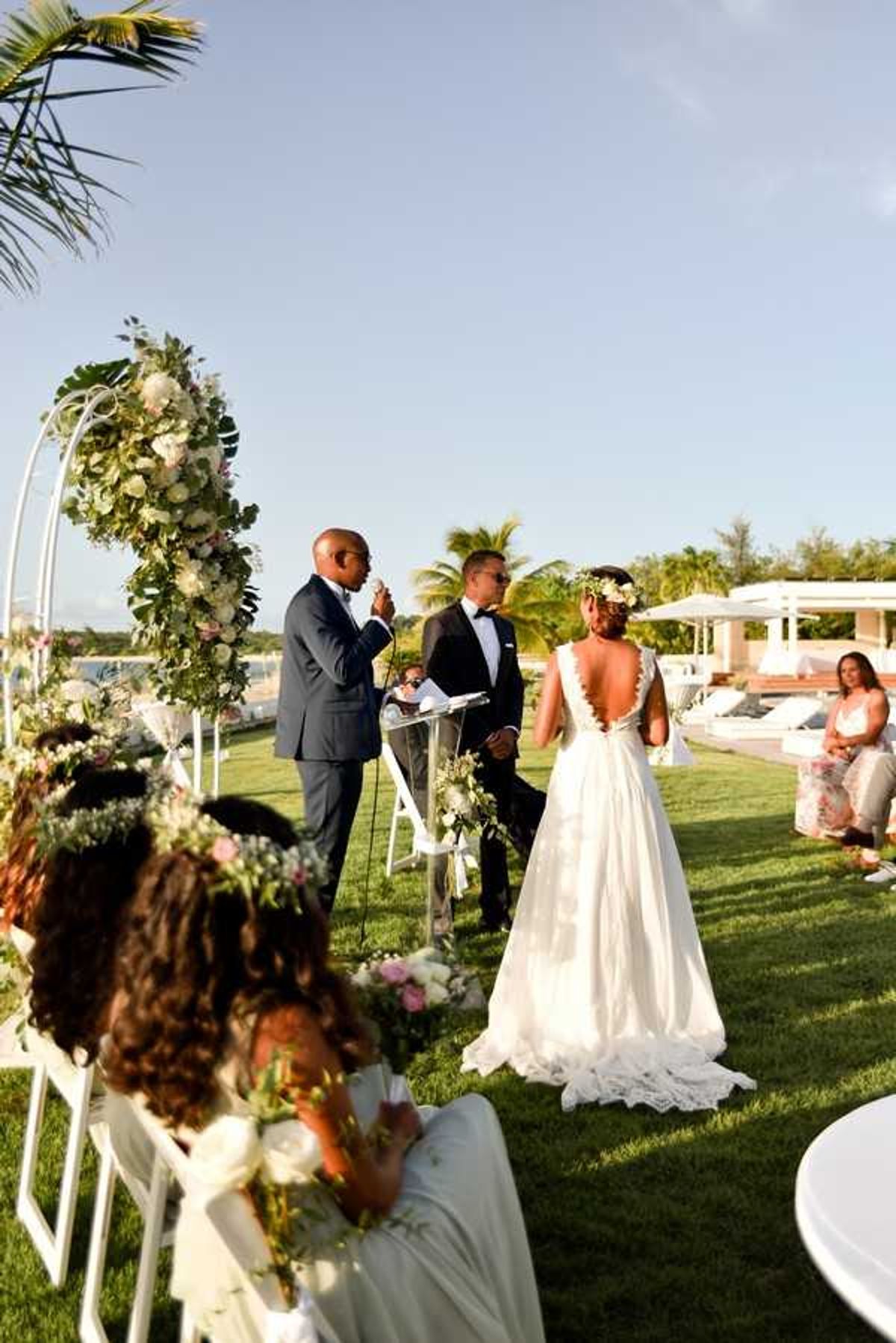 An outdoor wedding ceremony taking place on a manicured lawn in a tropical resort setting, with a pool area and white cabanas visible in the background. The bride wears a white gown with a deeply cut open back and lace detailing, paired with a floral crown of white and blush blooms, while the groom wears a black tuxedo with bow tie; an officiant in a navy suit speaks at a clear acrylic podium. The ceremony arch is a curved white metal structure adorned with a lush arrangement of white and blush roses, tropical monstera leaves, and greenery. In the foreground, two young flower girls in white dresses and floral crowns are seated in white folding chairs, holding small bouquets; the shot is taken from a medium-wide angle slightly behind and to the side of the guests.
