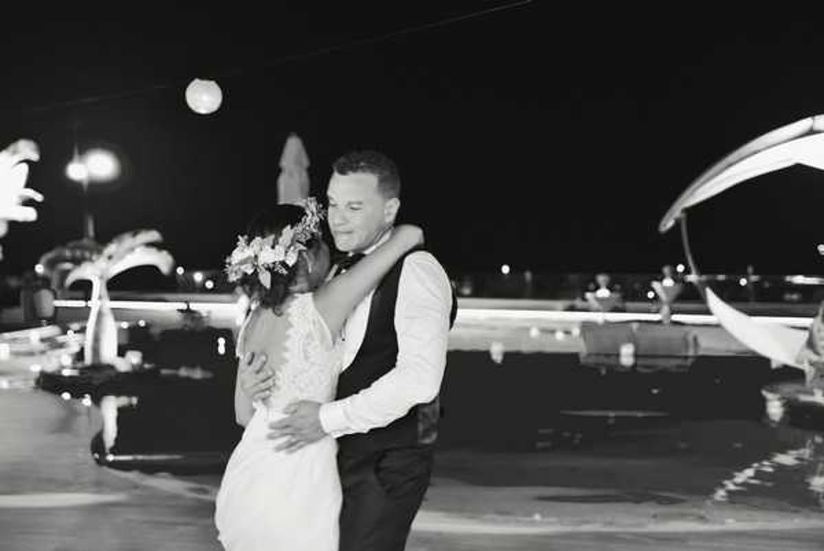 A black-and-white nighttime portrait of a couple sharing a close embrace, likely during a first dance or a quiet moment at an outdoor evening reception. The bride wears a fitted lace gown and has a floral hair piece, while the groom is dressed in a white dress shirt, dark vest, and bow tie. The background shows a poolside or waterfront terrace setting with scattered point-light sources — globe lights, lampposts, and bokeh reflections on water — creating strong contrast against the dark sky. The image is a mid-shot with soft focus on the background, drawing attention to the couple in the foreground.