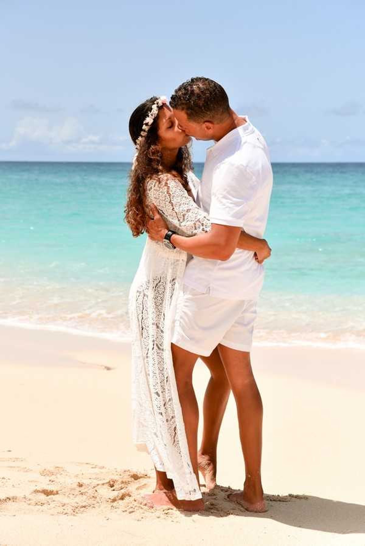 A couple shares a kiss during a beach portrait session, standing barefoot on white sand at the water's edge with turquoise ocean water visible behind them. The bride wears a long white crochet lace dress with three-quarter sleeves and a floral crown with small pale pink and white blooms in her curly hair. The groom is dressed in an all-white outfit consisting of a short-sleeve button-up shirt and shorts, wearing a dark watch. The styling is relaxed boho beach with an all-white color palette for both outfits. The shot is a full-length portrait taken in natural daylight.