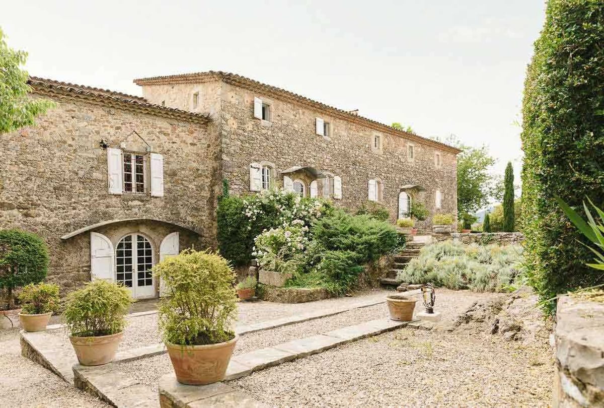 Wide exterior shot of a large Provençal stone farmhouse or mas, featuring traditional terracotta roof tiles, white-shuttered windows, and an arched double French door entrance. The foreground shows a gravel courtyard with terracotta pots planted with topiary shrubs and ornamental plants, along with stone steps leading up to a terraced garden area. White flowering shrubs are visible growing against the facade, and cypress trees frame the right side of the building. No people are present in the image. Potential venue feature image.