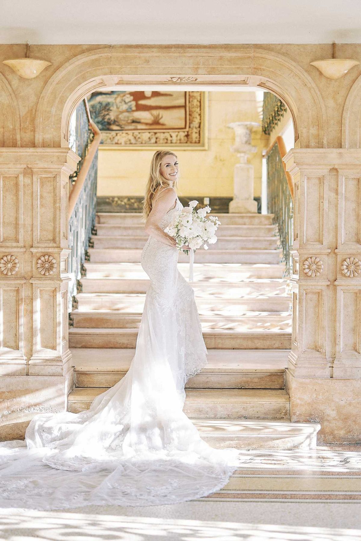 A bridal portrait taken indoors at the base of a grand marble staircase framed by an ornate carved stone archway with decorative rosette panels and wall sconces. The bride stands on the bottom step, turned slightly away from camera and glancing back with a smile, wearing a fitted lace mermaid-style gown with spaghetti straps and an extended lace train that spreads across the floor. She holds a rounded bouquet of white ranunculus and garden roses with a white ribbon. A large framed tapestry painting and wrought-iron balustrade are visible in the background through the arch. The image is a full-length portrait shot in bright, airy natural light. Potential venue feature image.