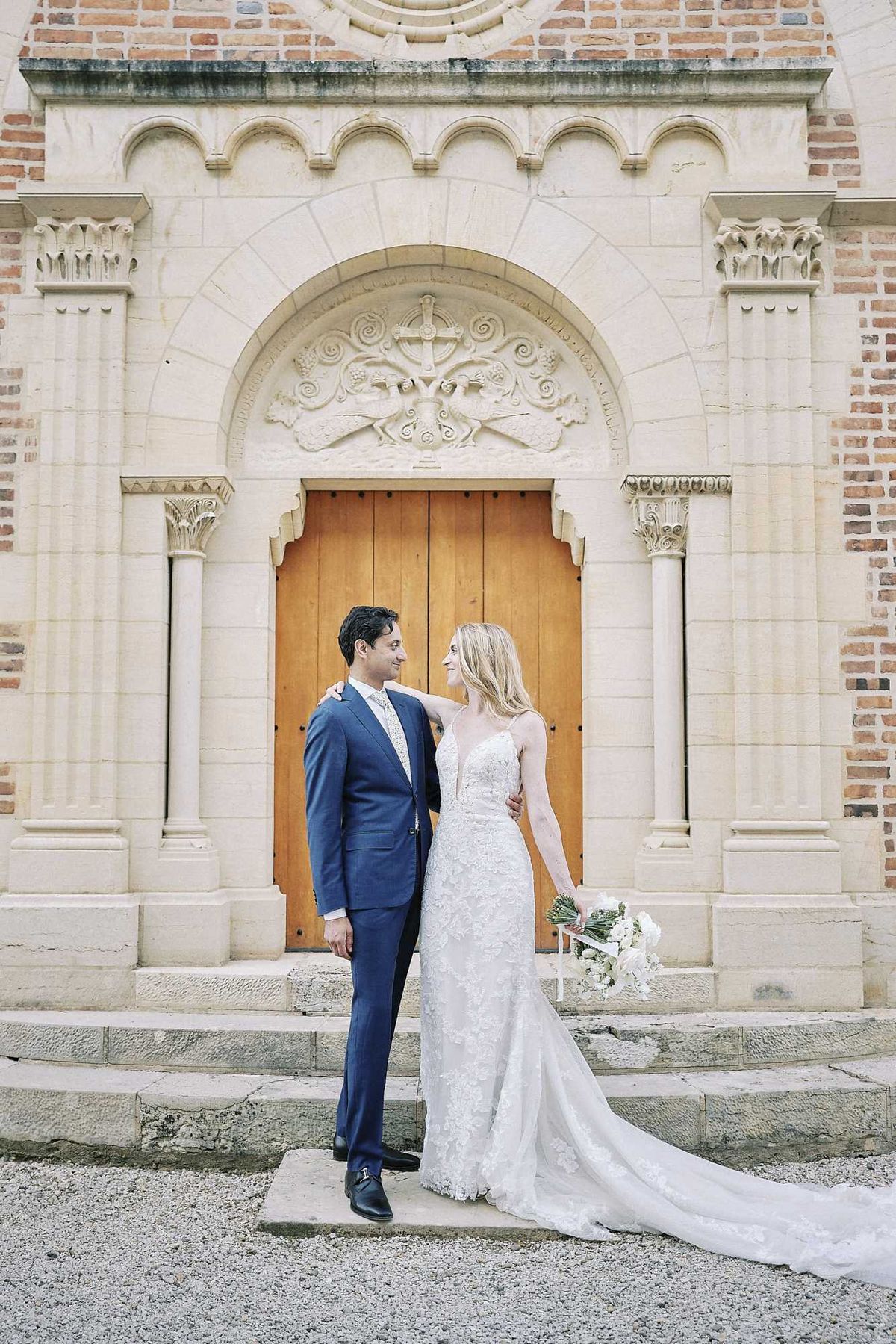 A couple portrait taken outdoors in front of an ornate chapel or church entrance featuring a carved stone archway with decorative relief work, flanked by stone columns, and large wooden double doors. The groom wears a navy blue suit with a light grey tie and black dress shoes, while the bride wears a fitted lace gown with spaghetti straps, a deep V-neckline, and a long train. She holds a bouquet of white flowers with greenery and has her arm around the groom's shoulder as they look at each other. The composition is a full-length couple portrait with the architectural facade as a prominent backdrop. Potential venue feature image.