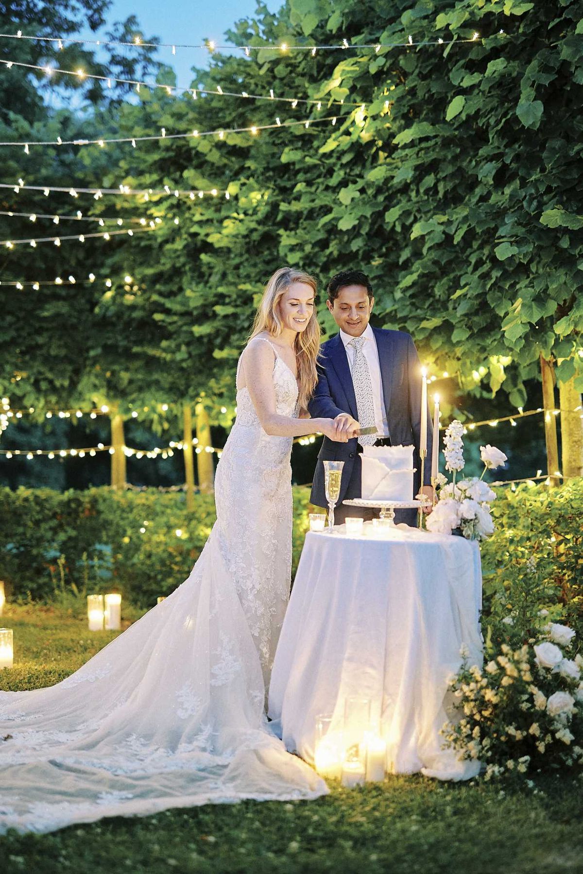 A bride and groom cut their wedding cake during an outdoor evening reception in a garden setting. The bride wears a fitted lace mermaid-style gown with a long train and thin straps, while the groom is dressed in a navy suit with a light grey patterned tie. The two-tier white cake sits on a white draped cocktail table accompanied by a champagne flute, tall tapered candles, and clusters of white roses and garden flowers at its base. Lighting is provided by strings of bistro fairy lights overhead and numerous pillar candles placed on the ground around the couple, creating a warm, intimate atmosphere against the dusk-blue sky. This is a medium full-length portrait shot of just the two of them.