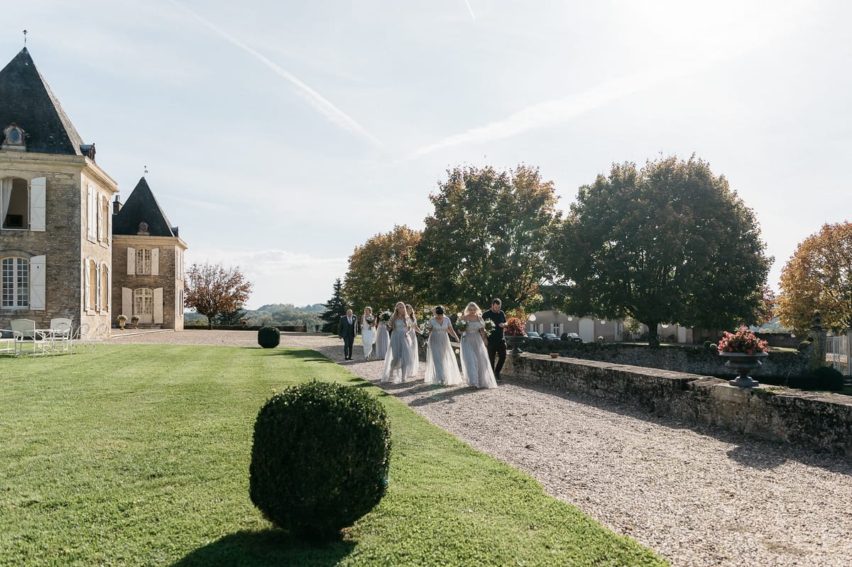 A bridal party processional captured in a wide outdoor shot along a gravel pathway at a French chateau. The group includes approximately four bridesmaids wearing floor-length dusty blue tulle gowns carrying white bouquets, accompanied by what appears to be a groomsman in a dark suit and additional members of the wedding party walking behind them. The chateau facade, featuring pointed slate rooftops, stone masonry, and white shuttered windows, is visible to the left, with a manicured lawn and clipped spherical boxwood topiary in the foreground. The styling is classic French country with a soft, romantic color palette centered on the pale blue bridesmaid dresses against the warm stone architecture. Potential venue feature image.