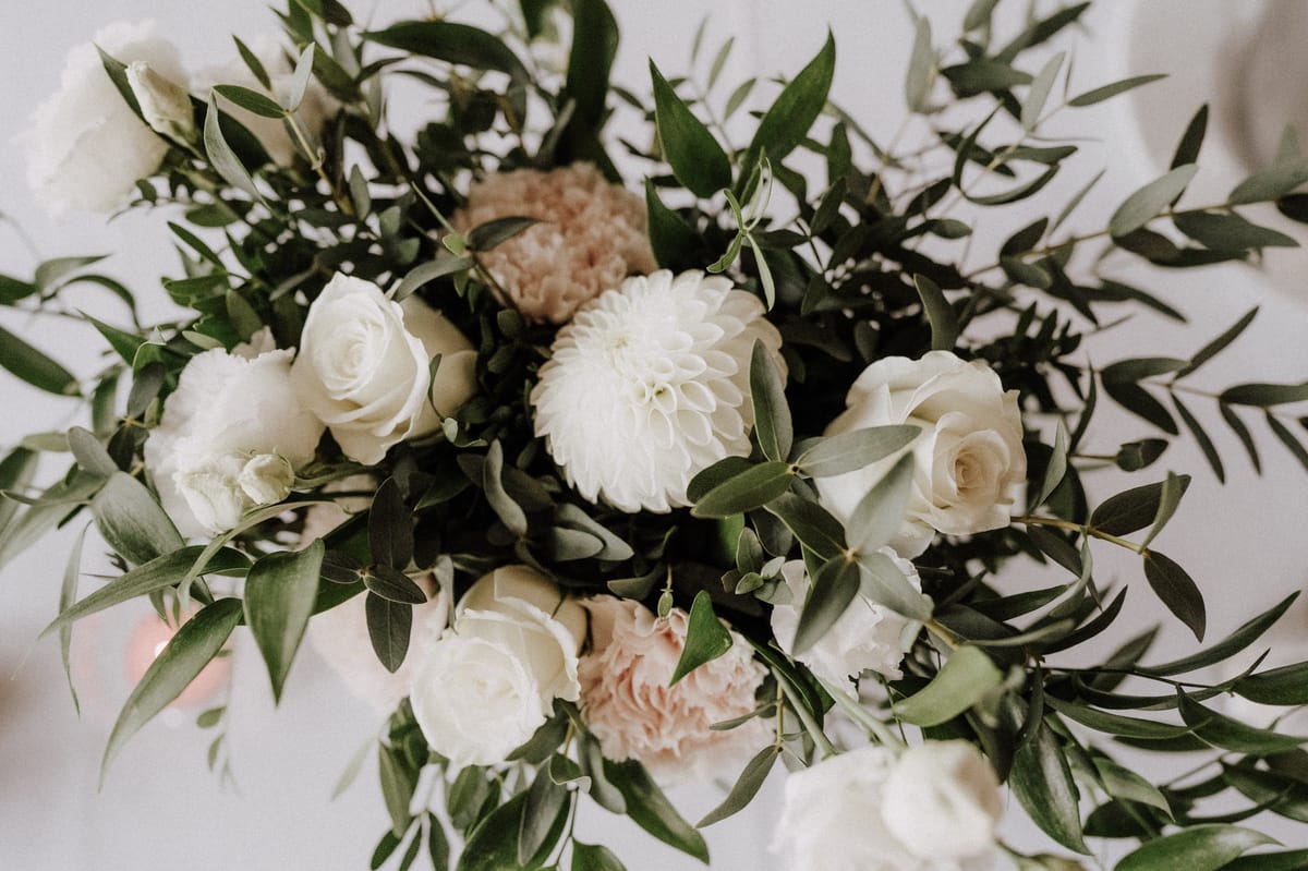 Close-up detail shot of a wedding floral arrangement featuring ivory roses, a white dahlia, white lisianthus, and blush carnations, interspersed with abundant loose olive branch foliage. The color palette is white, ivory, and soft blush against deep green, giving the arrangement a natural, organic feel consistent with a classic or garden-style aesthetic. The arrangement is photographed from above at a slight angle against a white surface, with a soft pink light source faintly visible in the lower left corner. No people are visible in the frame.