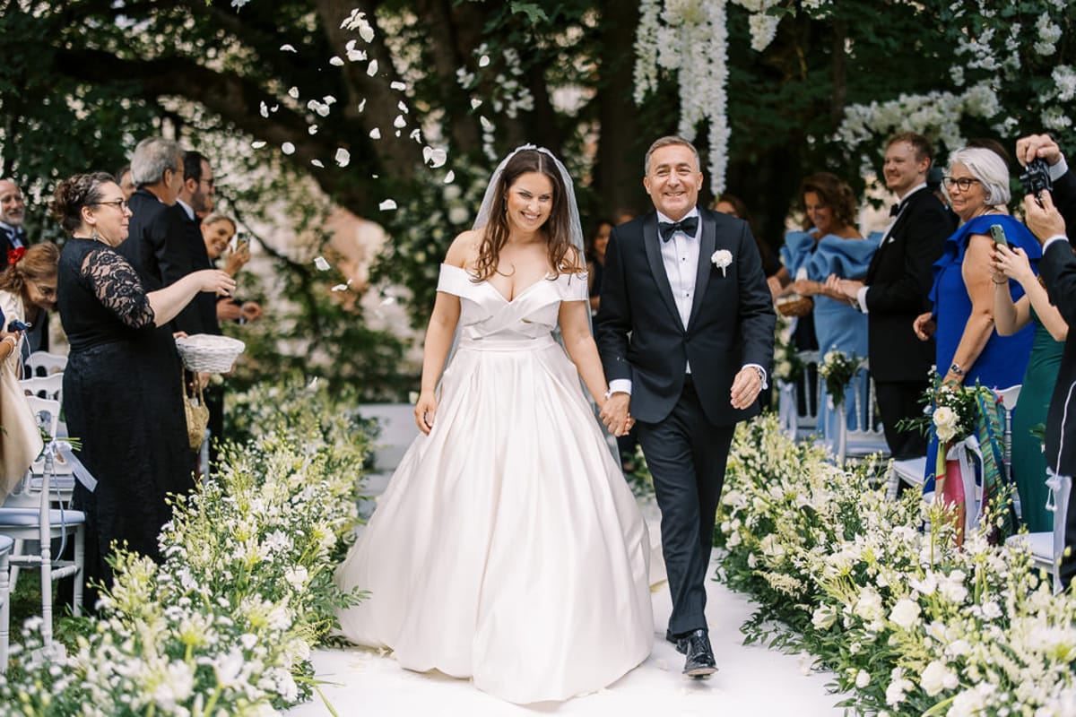 The bride and groom walk back down the aisle together following an outdoor ceremony, with guests on both sides tossing white flower petals over them. The bride wears an off-the-shoulder ivory ballgown with a full satin skirt and a cathedral-length veil, while the groom is dressed in a black tuxedo with a bow tie and a white boutonnière. The aisle is lined with lush arrangements of white flowers — including what appear to be white lisianthus, freesia, and greenery — laid directly on the ground, and a floral arch with hanging white blooms is visible in the background. The ceremony took place in a garden or chateau grounds setting, and approximately 20–30 guests are visible, some in formal attire including a woman in a cobalt blue dress; the mid-length shot captures the couple centered and smiling, with natural daylight illuminating the scene.