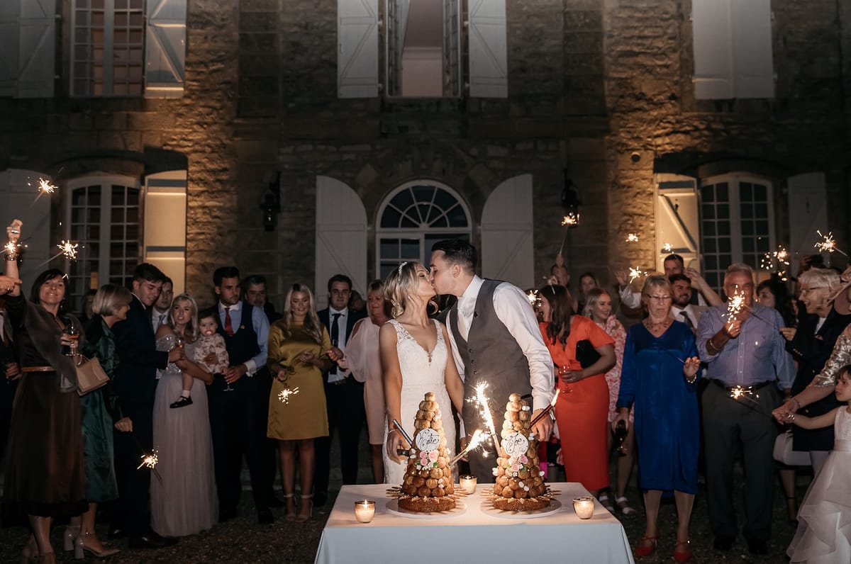 The bride and groom share a kiss during an outdoor croquembouche cutting ceremony at night, in front of a warmly uplighted chateau facade with arched windows and white shutters. Two croquembouche towers decorated with small pastel macarons and personalized calligraphy toppers sit on a white-clothed table in the foreground, each fitted with lit sparklers, alongside small votive candles. Approximately 30–40 guests form a semicircle around the couple, the majority holding lit sparklers aloft, creating a bright, celebratory atmosphere. The bride wears a lace V-neck fitted gown with a delicate hairpiece, while the groom is dressed in a white shirt and grey waistcoat; guests are dressed in a mix of formal attire including mustard yellow, teal, orange, and cobalt blue outfits. The wide-angle shot captures both the couple and the full crowd, with the illuminated chateau building prominently framing the scene in the background.
