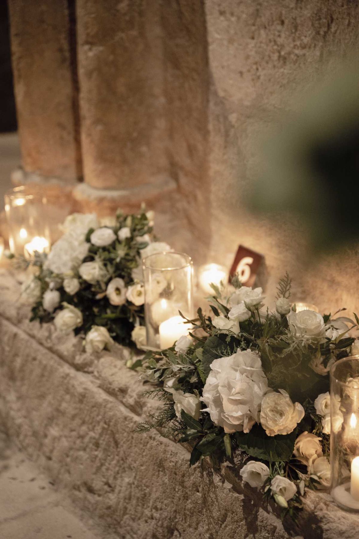 Close-up detail shot of wedding reception decor arranged along a low stone ledge at the base of stone columns, likely inside a historic venue such as a chapel or chateau. The arrangement features multiple clusters of white peonies, white roses, white ranunculus, and lisianthus with deep green foliage including ivy and fern. Glass cylinder candle holders and small votive candles are scattered throughout, casting warm candlelight across the stone surface. A dark red table number card displaying the number 6 is visible among the florals. The overall decor palette is white and green with warm candlelight, styled in a classic, romantic aesthetic.