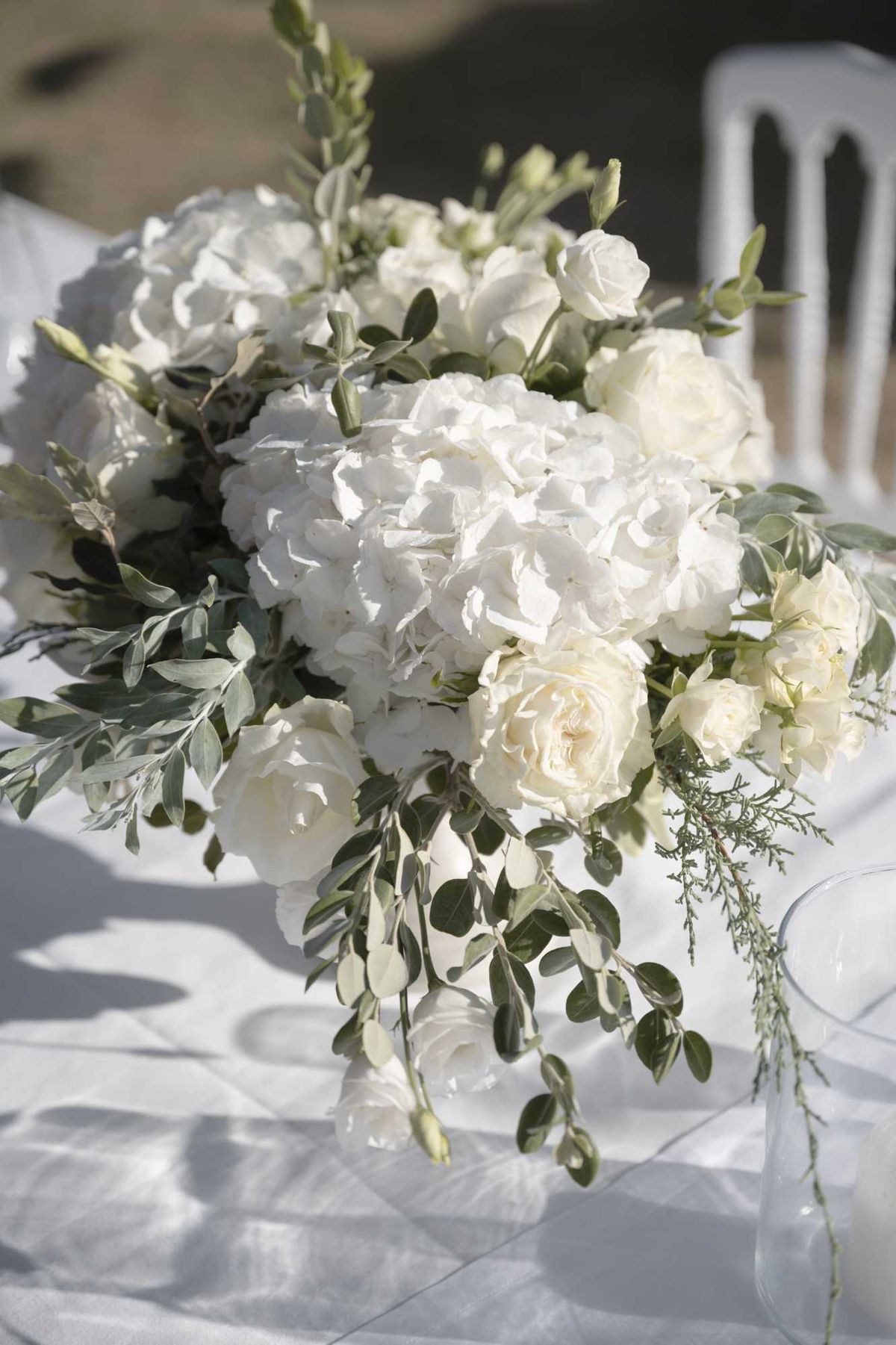 Close-up detail shot of an outdoor wedding table centerpiece arrangement placed on a white linen-covered table. The arrangement features white hydrangeas, cream garden roses, white spray roses, and white freesias, accented with trailing olive branches and wispy rosemary sprigs in a clear glass vase. The overall color palette is all-white and cream with green foliage, consistent with a classic or garden-style aesthetic. A white Napoleon chair is partially visible in the blurred background.