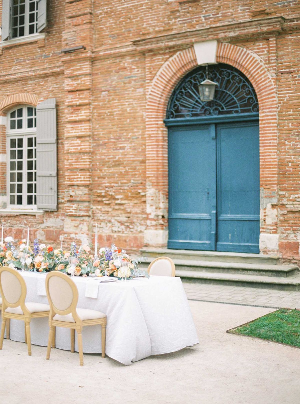 An outdoor reception tablescape is set in the courtyard of a French chateau, positioned directly in front of a large arched doorway with a distinctive deep blue double door and ornate wrought-iron fanlight. The rectangular table is covered with a white linen and lined with gold-framed Louis XVI-style chairs with cream upholstery. A lush floral runner stretches the length of the table, featuring peach and apricot roses, ivory blooms, lavender-toned flowers, and eucalyptus greenery, accompanied by tall white taper candles. The wide shot captures both the table styling and the red brick facade of the historic building, emphasizing the classic French architectural backdrop. Potential venue feature image.