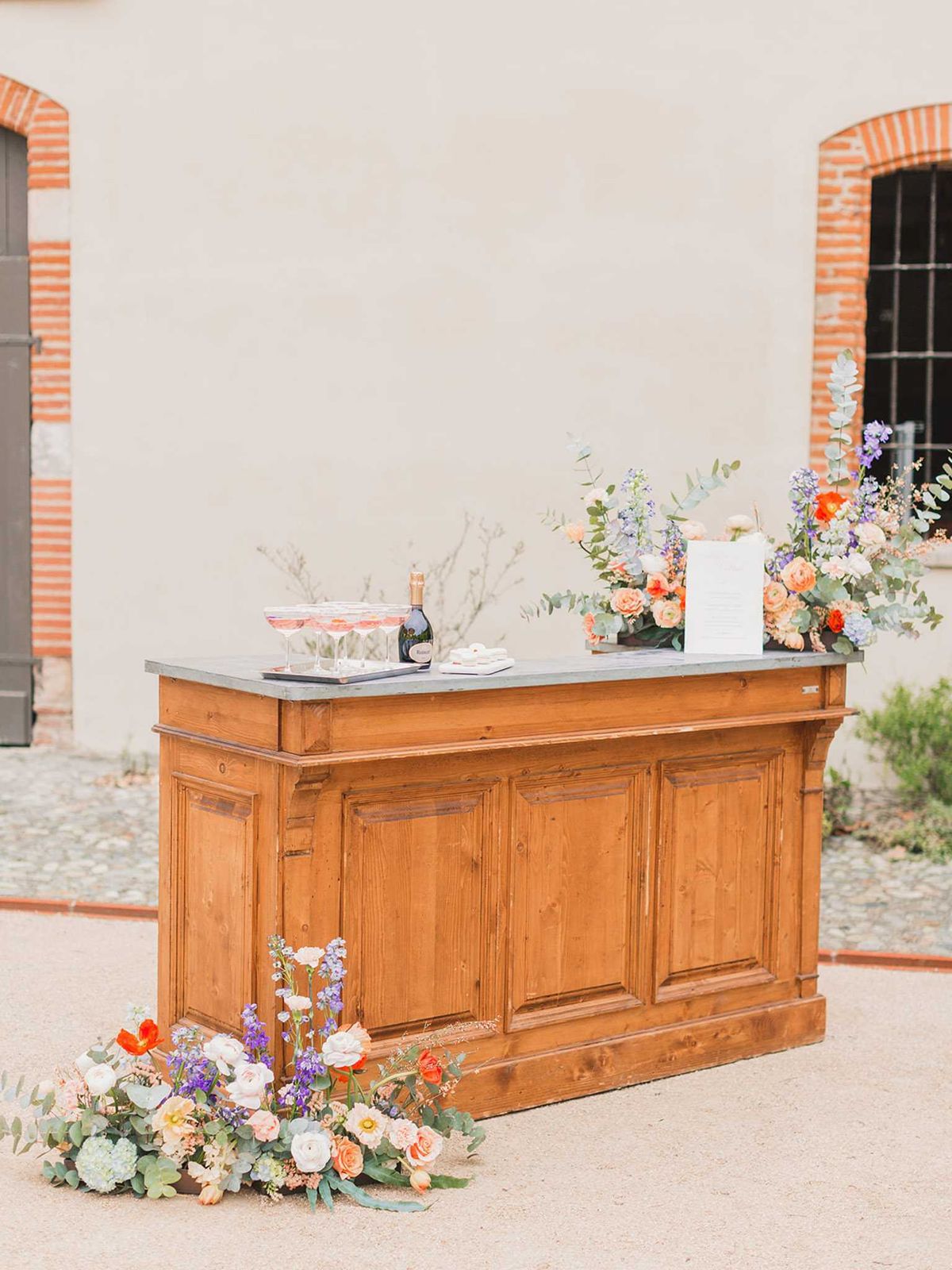 A cocktail hour bar station set up outdoors against the rendered facade of a French venue with red brick window surrounds. The bar is a vintage-style warm pine wood counter with a zinc or slate grey top, styled with two large floral arrangements featuring peach garden roses, orange ranunculus, lavender-blue delphiniums, white poppies, and eucalyptus foliage. A tray of pink champagne coupe glasses and a dark bottle of sparkling wine sit on the bar surface alongside a printed menu card. A ground-level floral arrangement at the base of the bar mirrors the same color palette of peach, orange, white, and purple blooms with soft green hydrangea and eucalyptus. The overall decor palette is a warm peach and coral paired with dusty purple and sage green, giving a garden-style aesthetic. Wide shot, no people present.