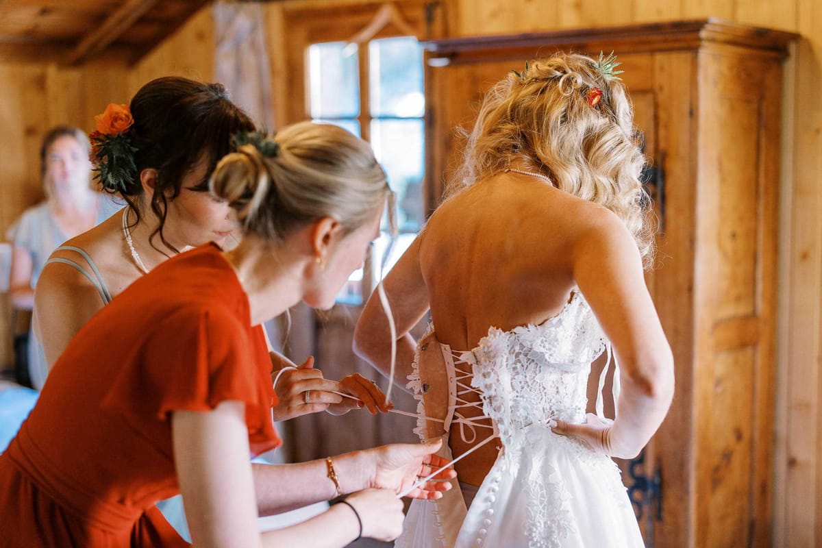 A getting-ready scene captured indoors in a warm-toned wooden chalet or cabin room, with a large dark wood armoire visible in the background. Two women are lacing up the back of the bride's white lace corset-back wedding dress — one in a burnt orange short-sleeve dress and one in a sage green top, both with updo hairstyles adorned with orange flowers and dark foliage. The bride, seen from behind, has loose blonde waves with small orange floral and greenery hair accessories, and wears a backless lace gown with ribbon lacing and pearl button details. A third woman is visible out of focus in the background. The overall styling palette is warm, with terracotta and orange tones carrying through the bridesmaid attire and hair florals. Medium close-up shot focused on the back of the dress.